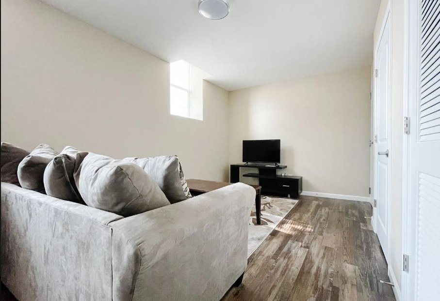 Living room with a beige sofa and pillows, small flat-screen TV on black stand, hardwood floors, white walls, small window, white closet doors.