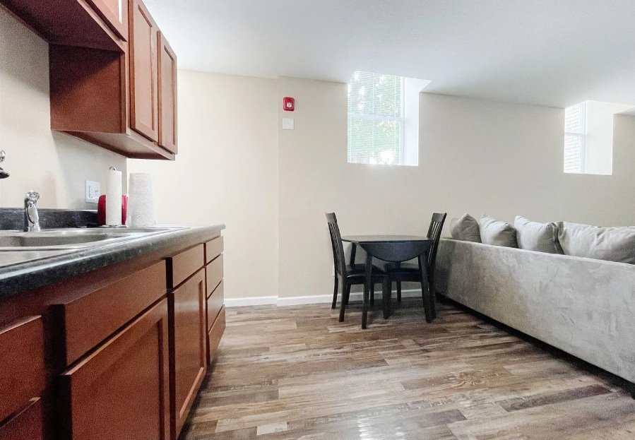 Living room with kitchenette, small round dining table with four chairs, beige sofa, two small windows, wood flooring, and beige walls.