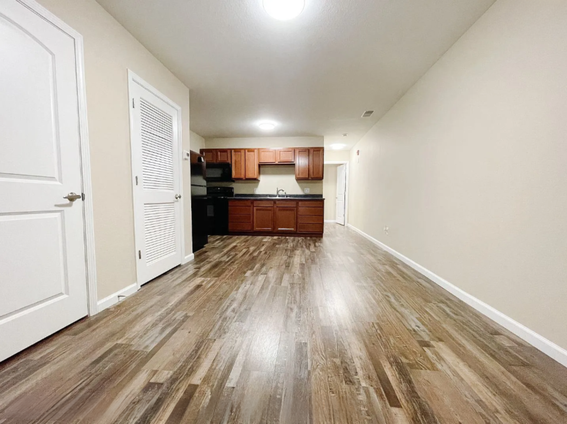 Empty living area with hardwood floors, white walls, and a kitchen with wooden cabinets and black appliances in the background
