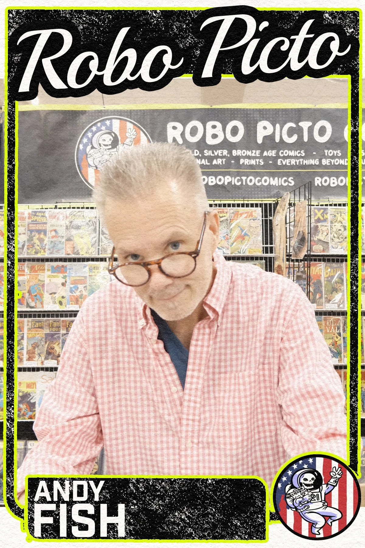 A man with gray hair and glasses wearing a pink checkered shirt, standing in front of a comic book booth called Robo Picto, at a comic convention, with visible comic books on shelves behind him.