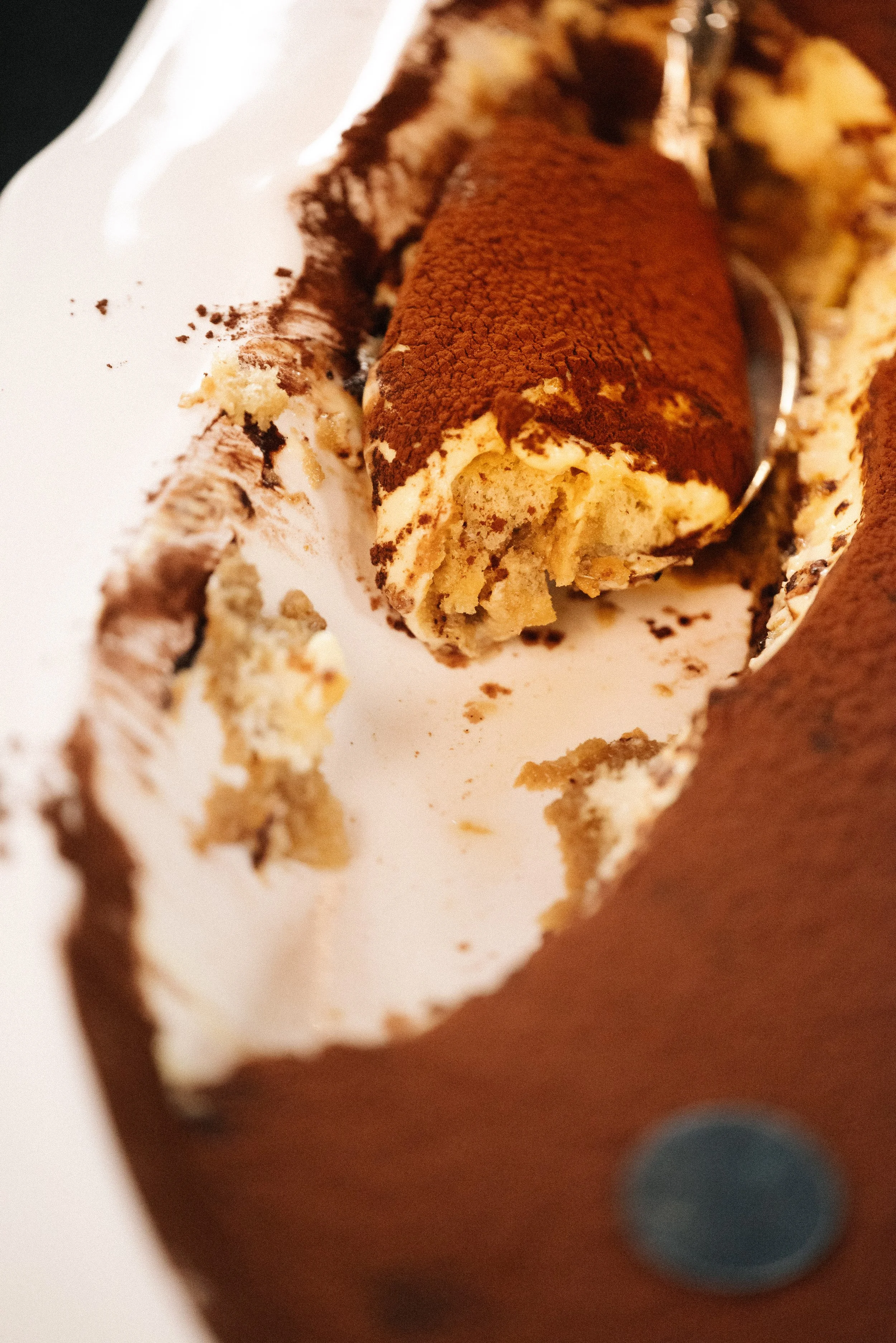 A partially eaten traditional Italian tiramisu dessert, showing layers of coffee-soaked sponge cake, mascarpone cheese, and cocoa powder on top, served on a white plate.