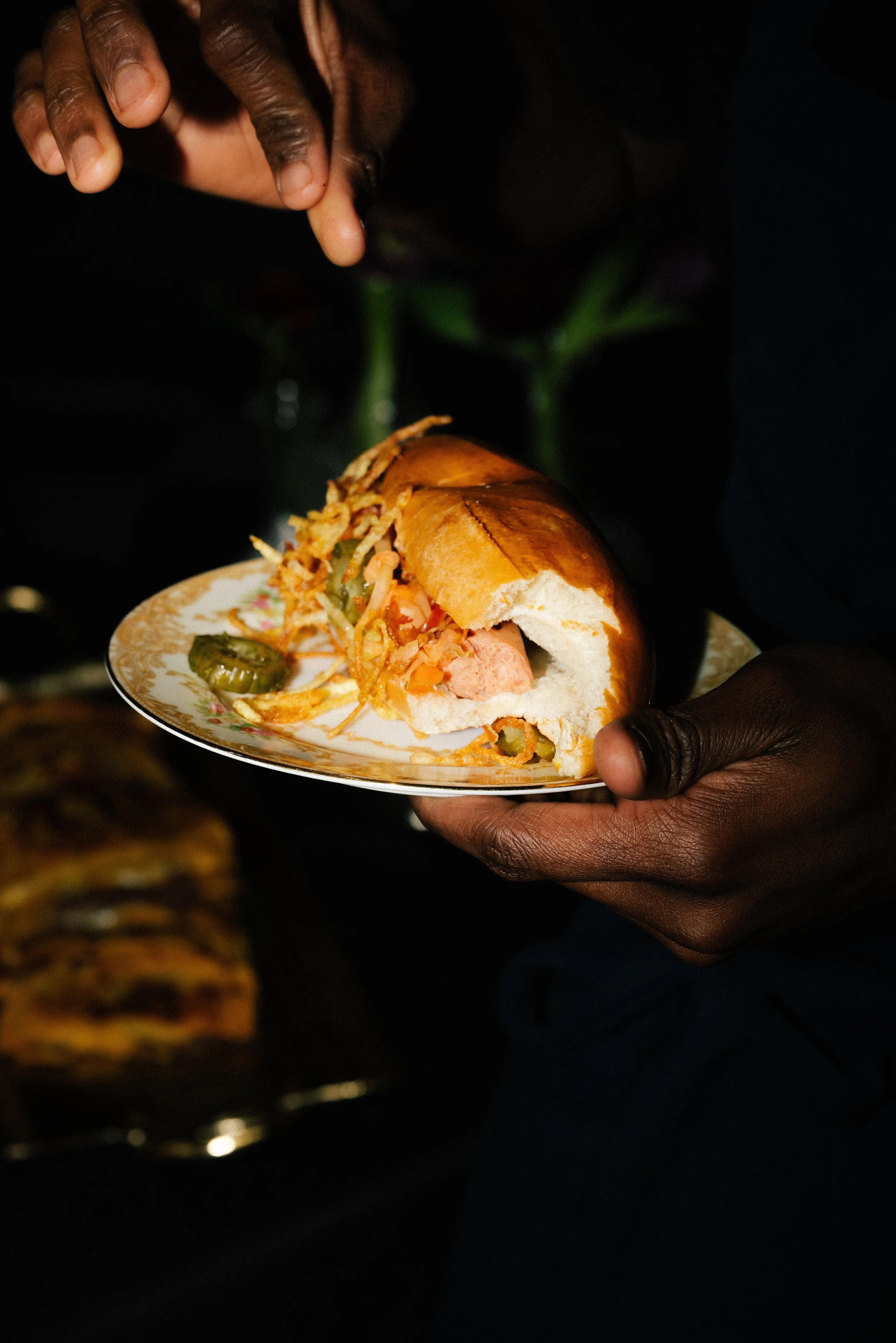 A person holding a plate with a sandwich, featuring some pickles and coleslaw, in a dark setting.