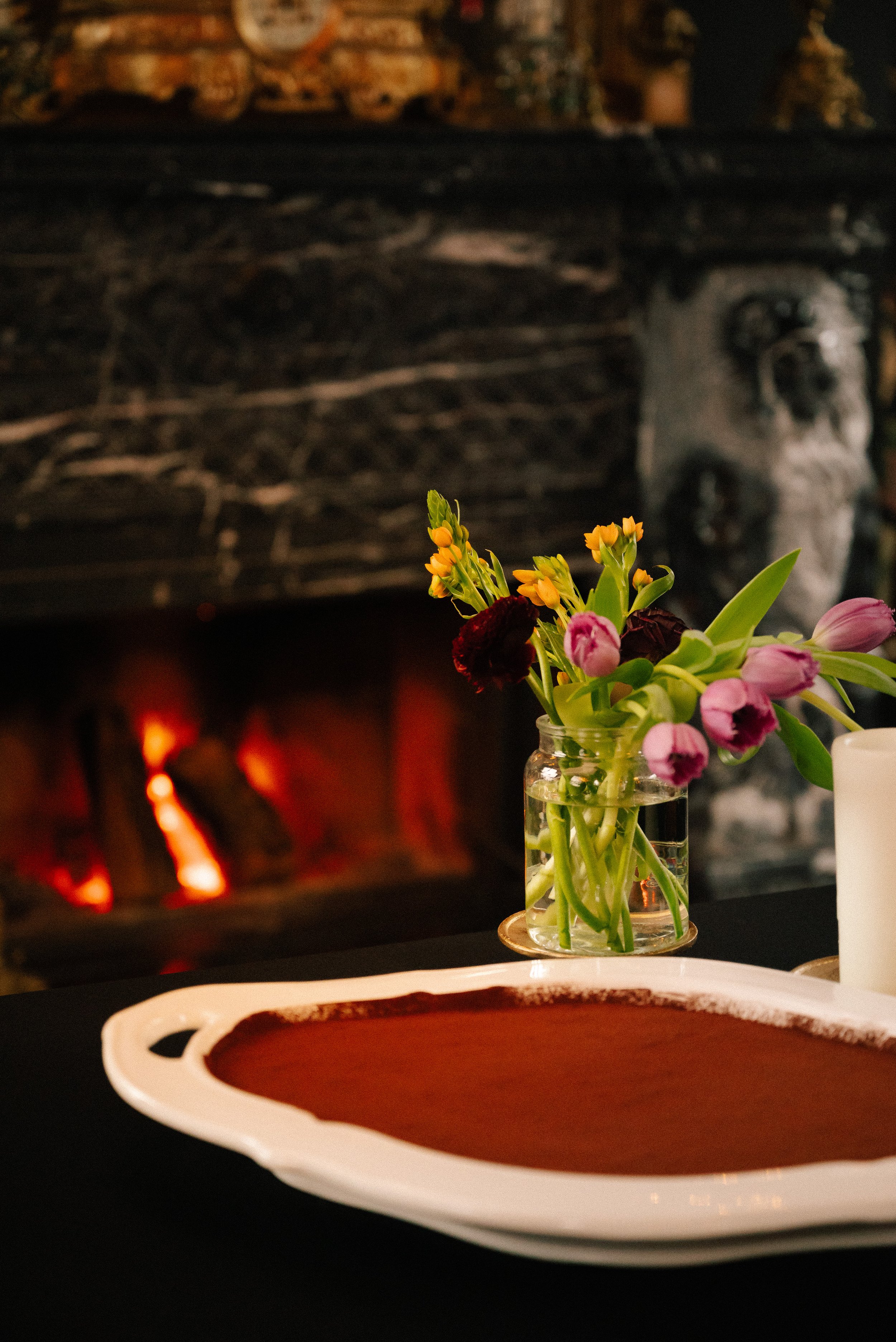 A bouquet of colorful flowers in a glass vase on a table in front of a fireplace with glowing logs.