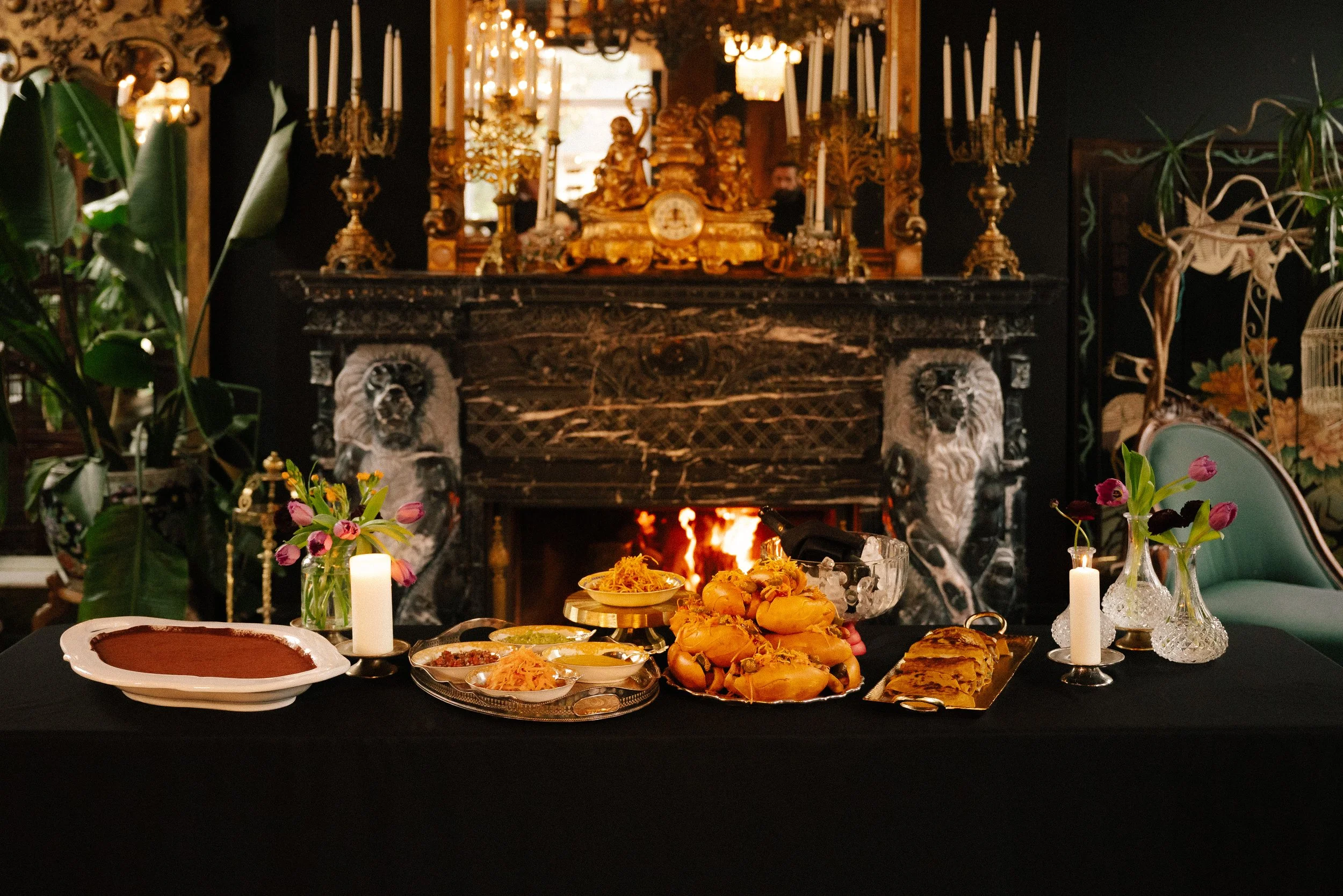 A table set for a meal in front of a fireplace with ornate decorations, candles, plants, and flowers, suggesting a festive or elegant occasion.