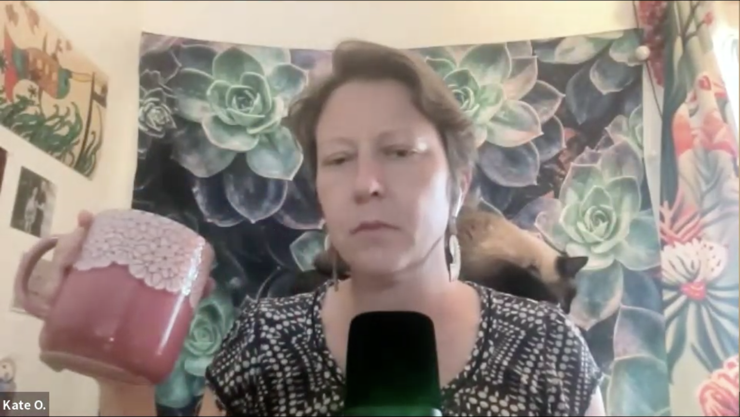 Kate sits at her desk holding a giant pink teacup. A cat rests on the top of her chair behind her head.