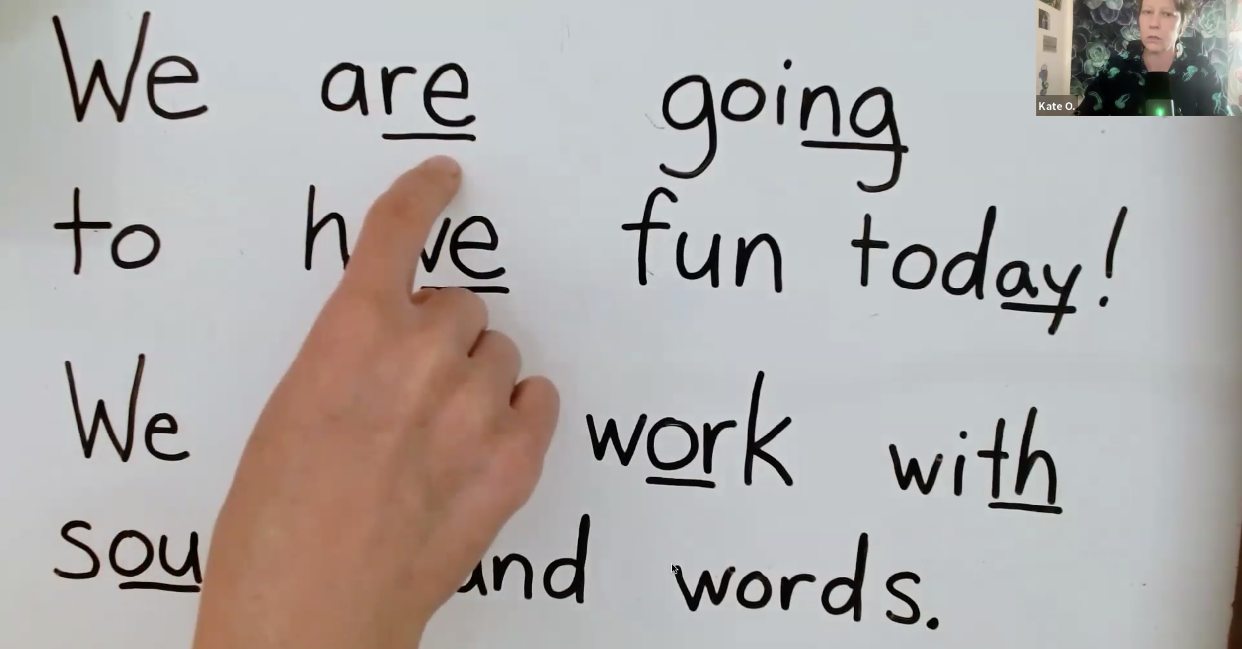 Kate points to a whiteboard with lines under specific words that analyze spelling patterns. The board reads, We are going to have fun today! We will work with sounds and words.