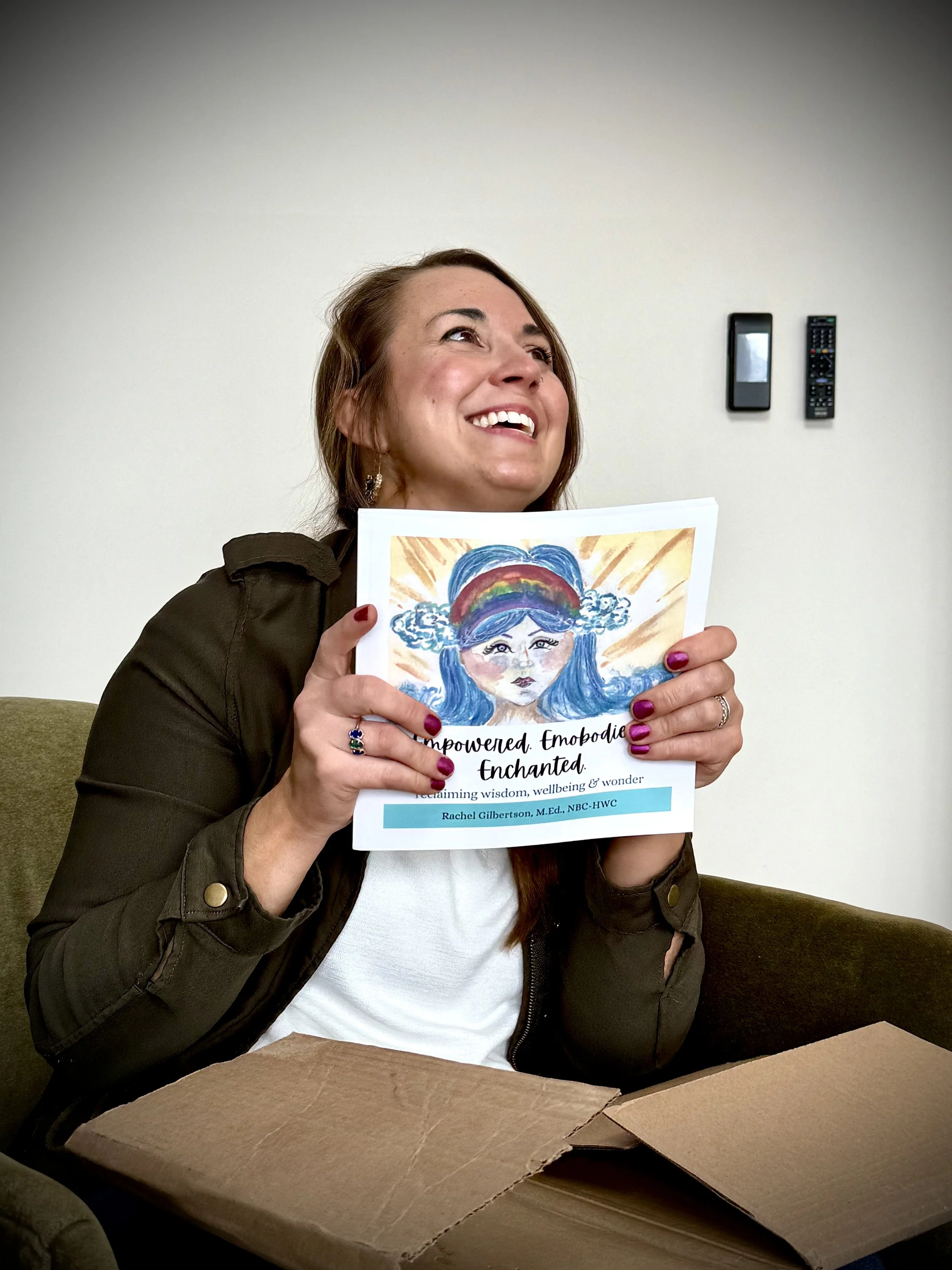 A woman with brown hair sitting on a green couch, smiling and holding up a colorful illustrated booklet titled 'Empowered, Emobodie, Enchanted' with a rainbow-haired character. She has painted nails and wears a dark jacket.