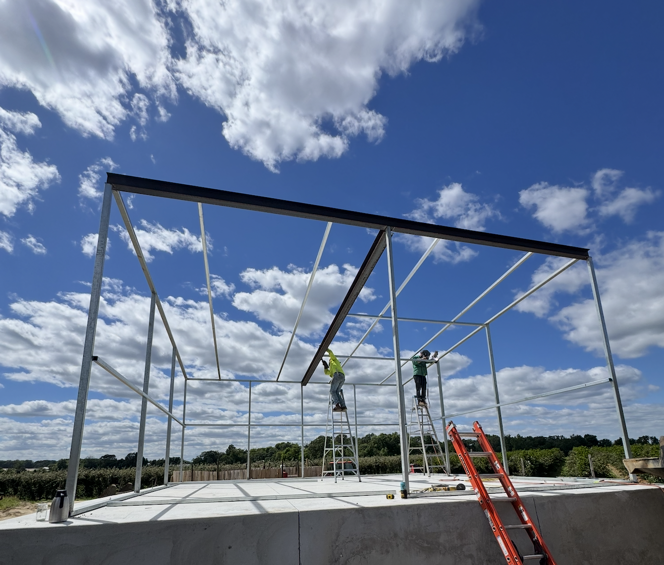 Two workers on ladders assembling the frame of a building outdoors on a partly cloudy day.
