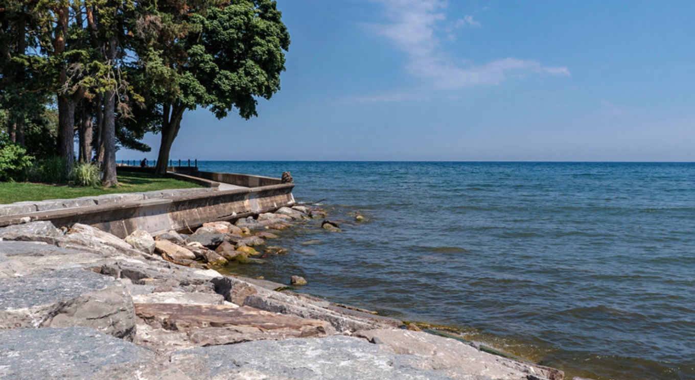 View of a lakeside with a stone and concrete walkway, large trees, and calm water under a partly cloudy sky.