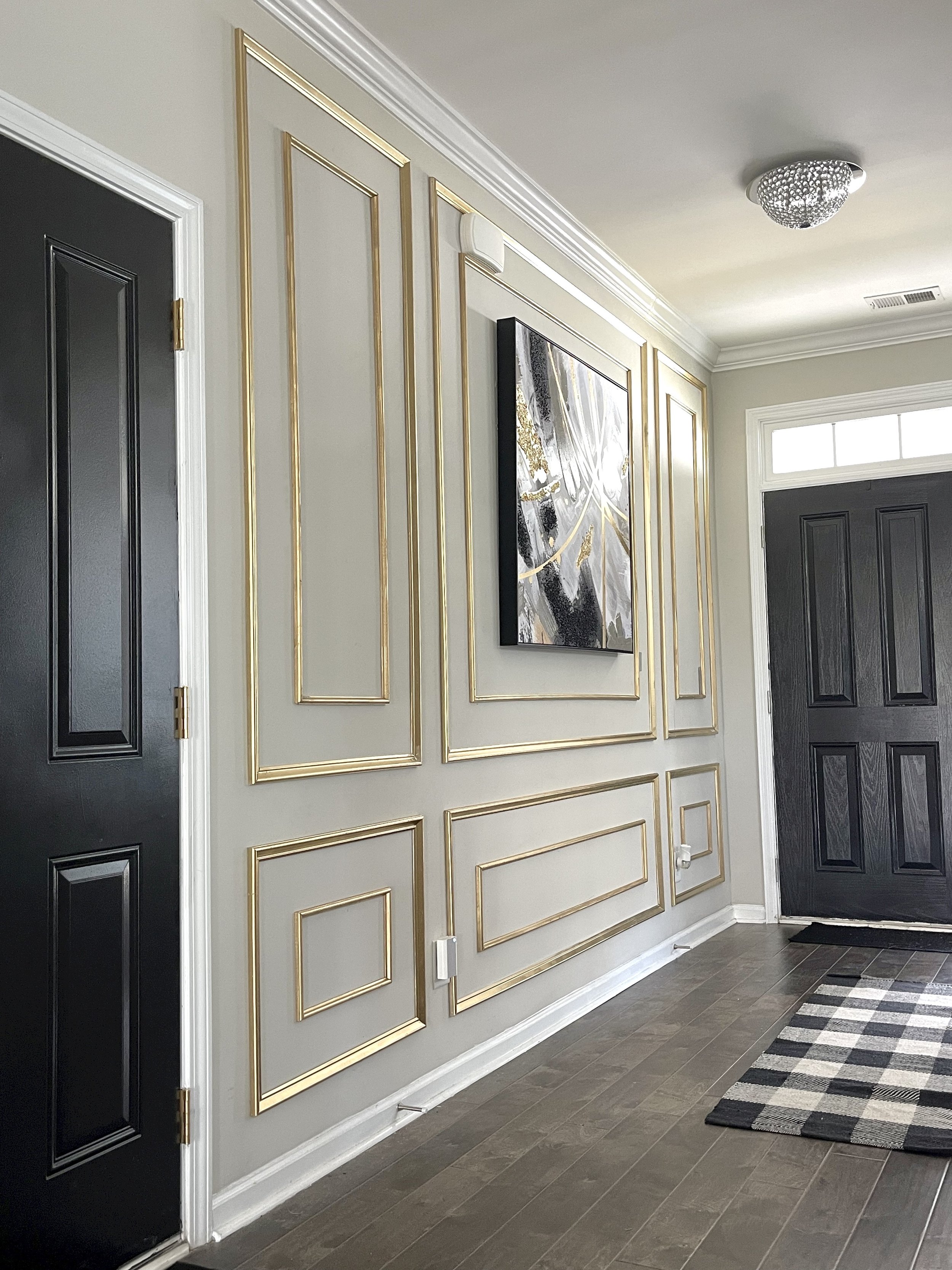 Interior of a modern entryway with beige walls decorated with gold trim, black doors, a black and white checkered rug, a framed abstract painting, and a ceiling light fixture.