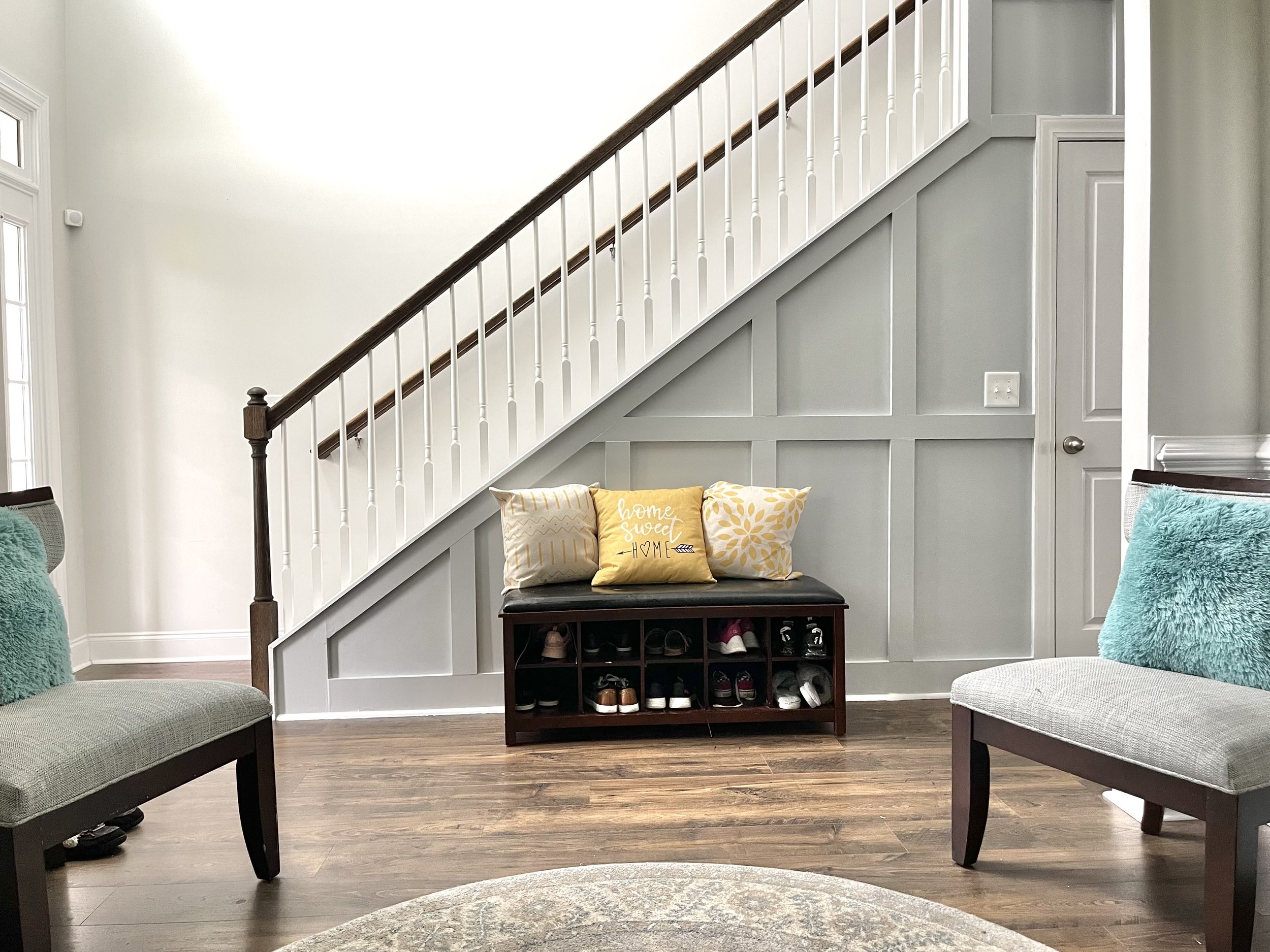Living room with a staircase featuring white railings and dark wood handrail, a small shoe-storage bench with decorative pillows, and two chairs with teal fuzzy cushions, a light-colored area rug, and hardwood floors.