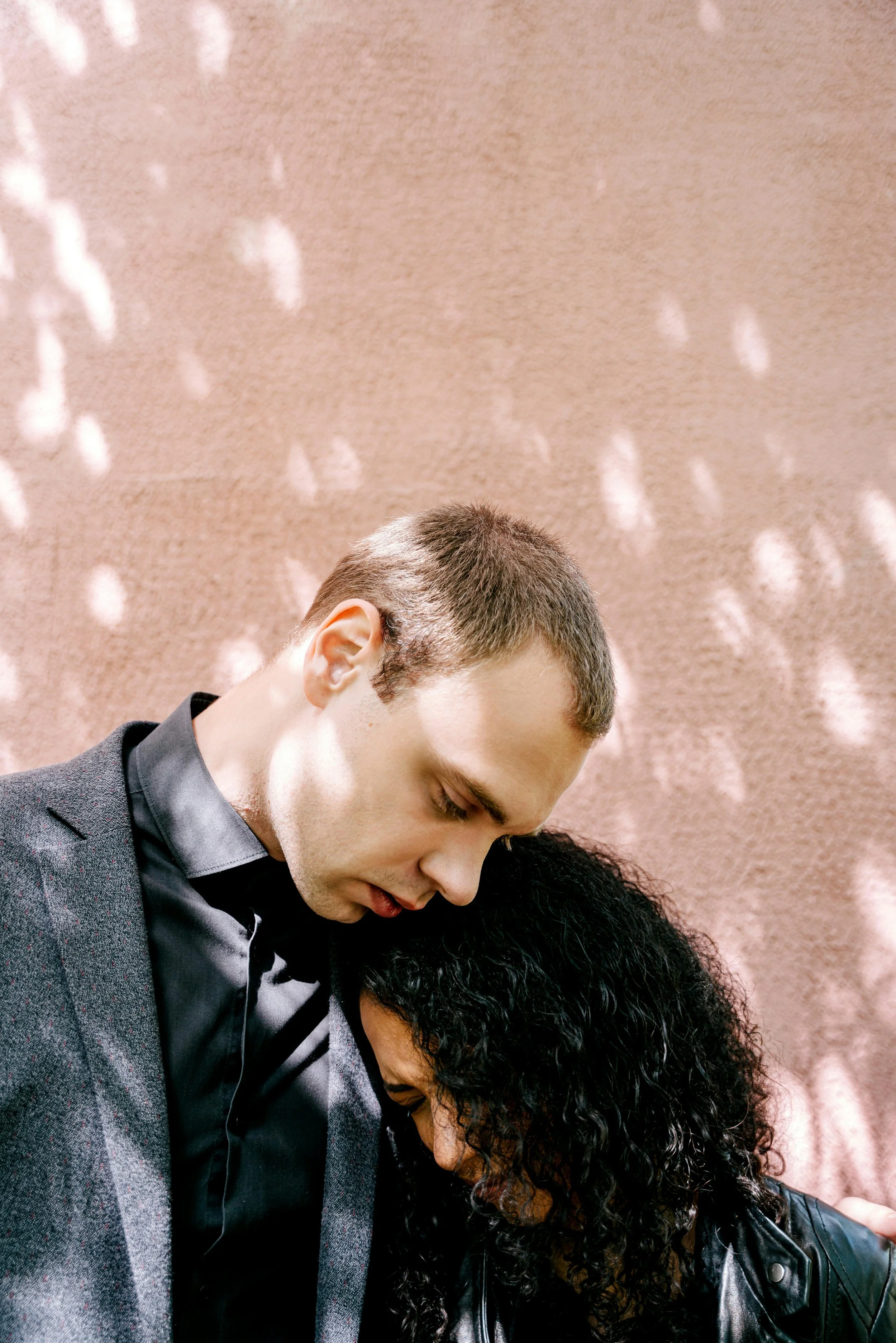 A man and a woman with their foreheads touching, head bowed, and eyes closed. The man has short brown hair, is wearing a dark suit and shirt. The woman has curly black hair and is wearing a black leather jacket. They are standing against a beige back