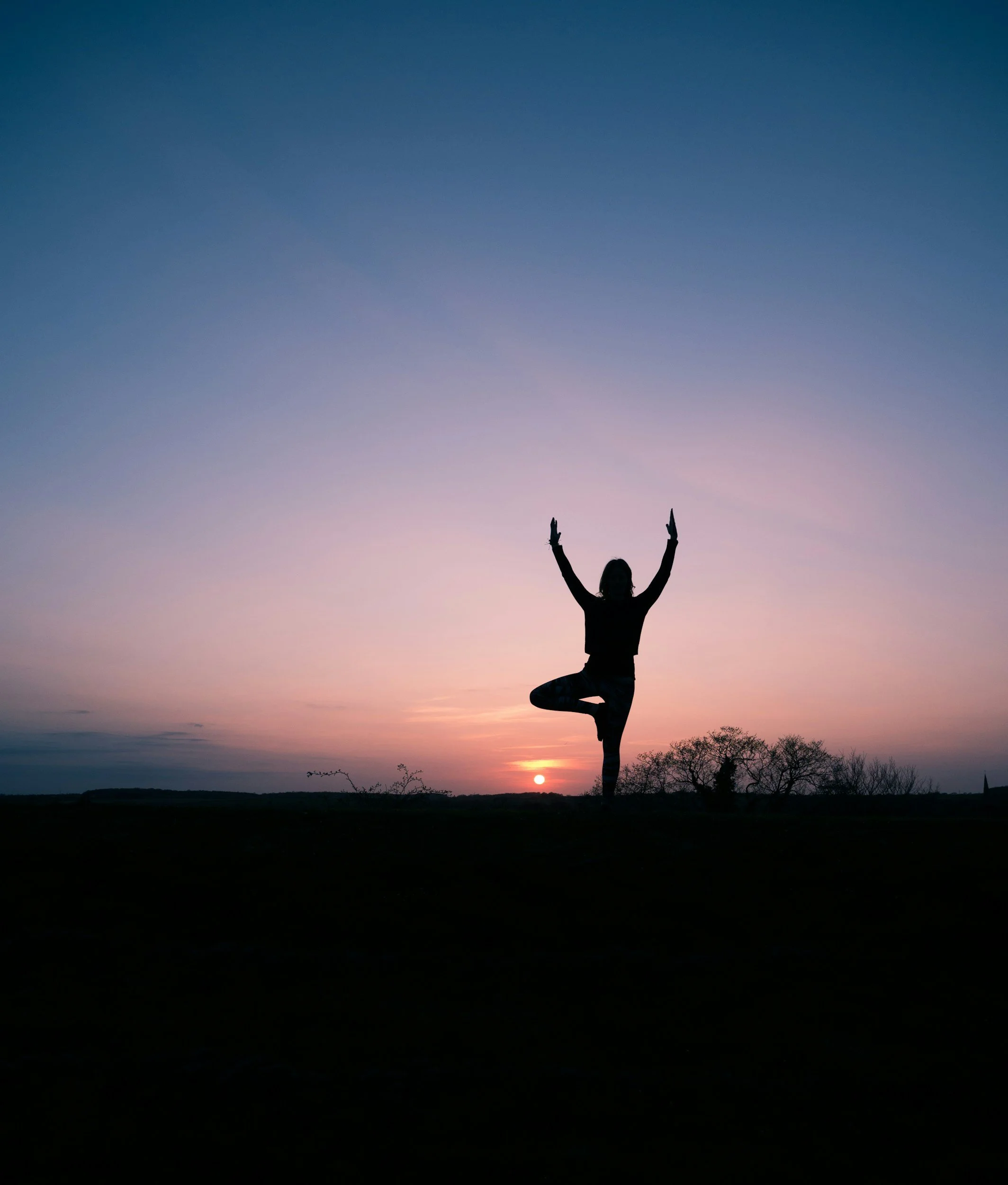 Silhouette of a person practicing yoga in a tree pose during sunset, with the sun near the horizon and a colorful sky.