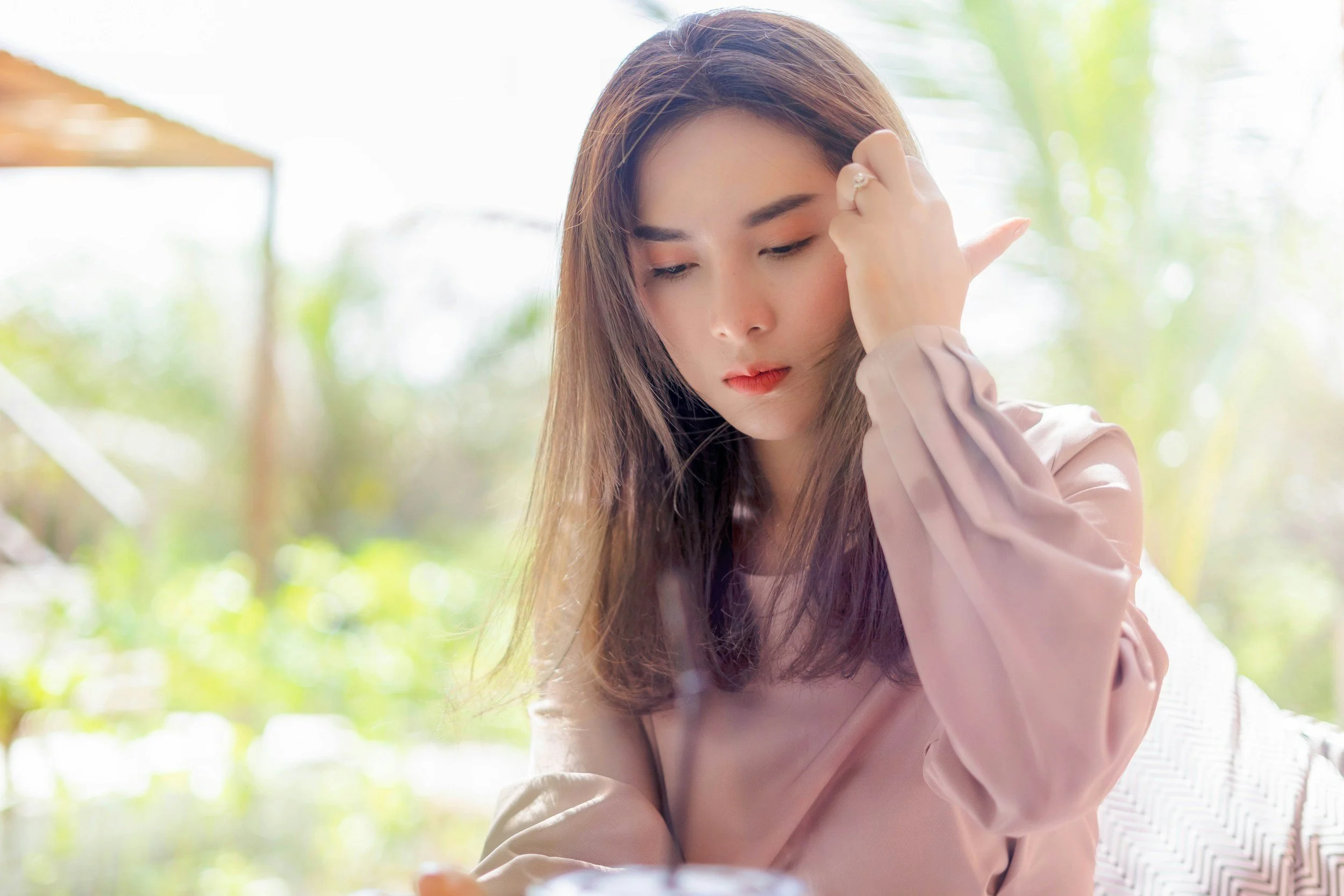 A young woman with long brown hair looking down thoughtfully, with a pensive expression, sitting outdoors with blurred green foliage in the background.
