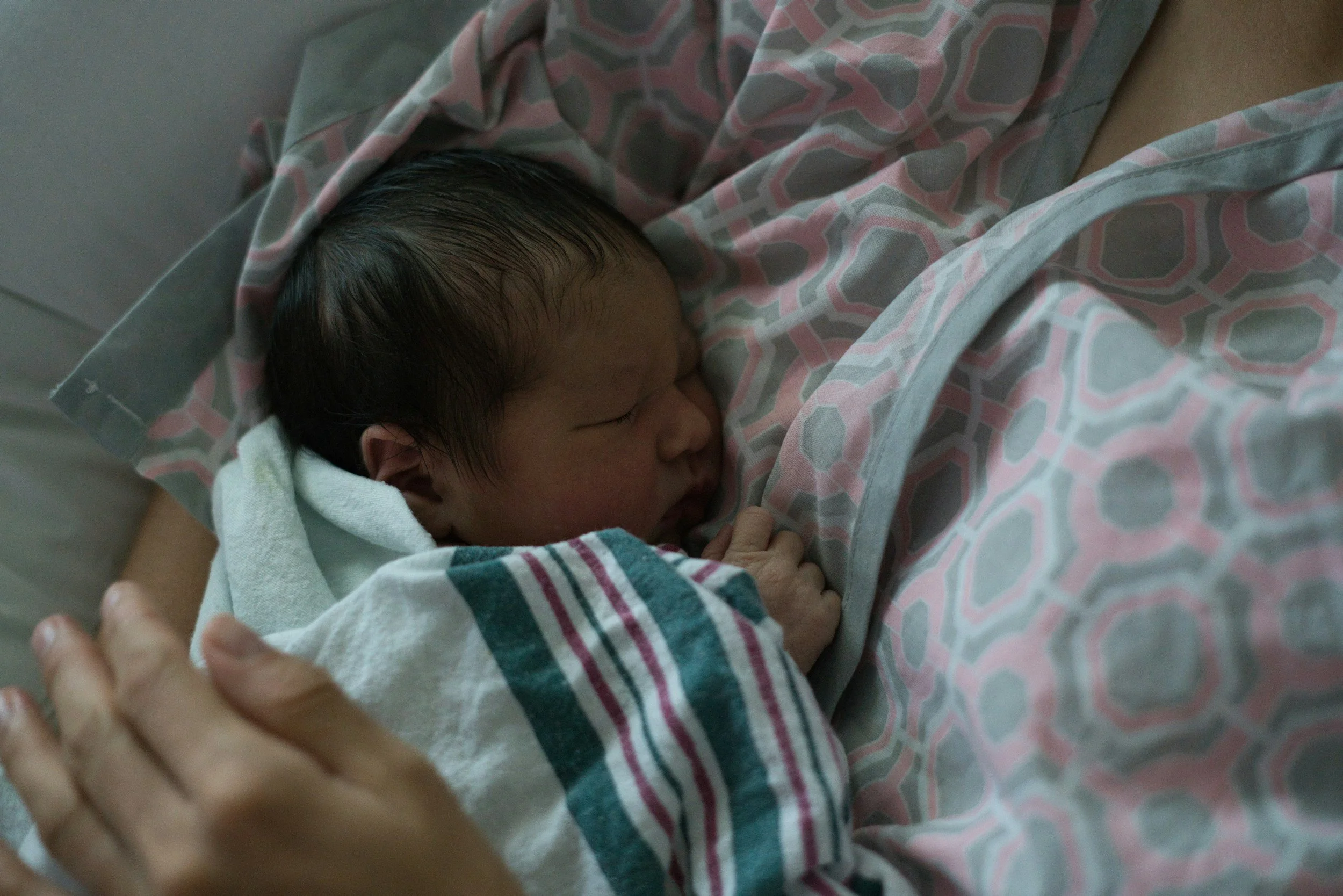 A newborn baby with dark hair sleeping, wrapped in a hospital blanket with pink and gray geometric patterns, lying on a hospital bed.