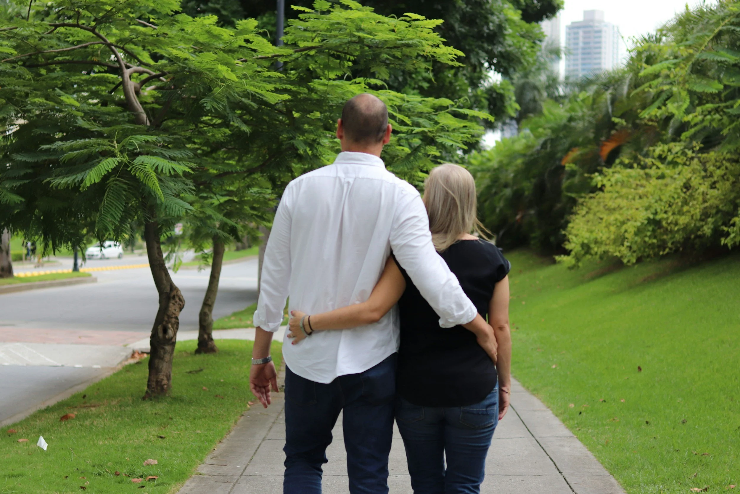 A couple walking arm in arm along a park sidewalk surrounded by green trees and grass, with city buildings visible in the background.