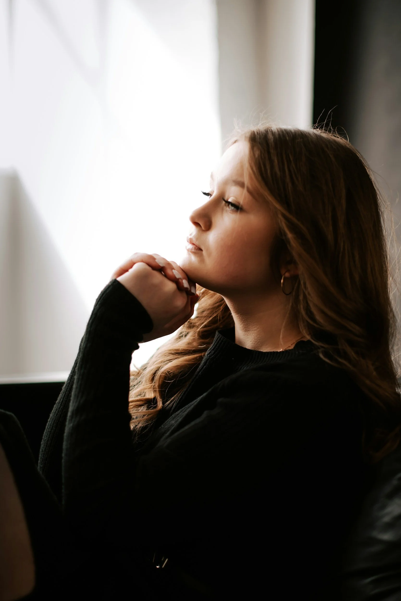 A young woman with long, wavy brown hair sitting near a bright window, resting her chin on her hand.