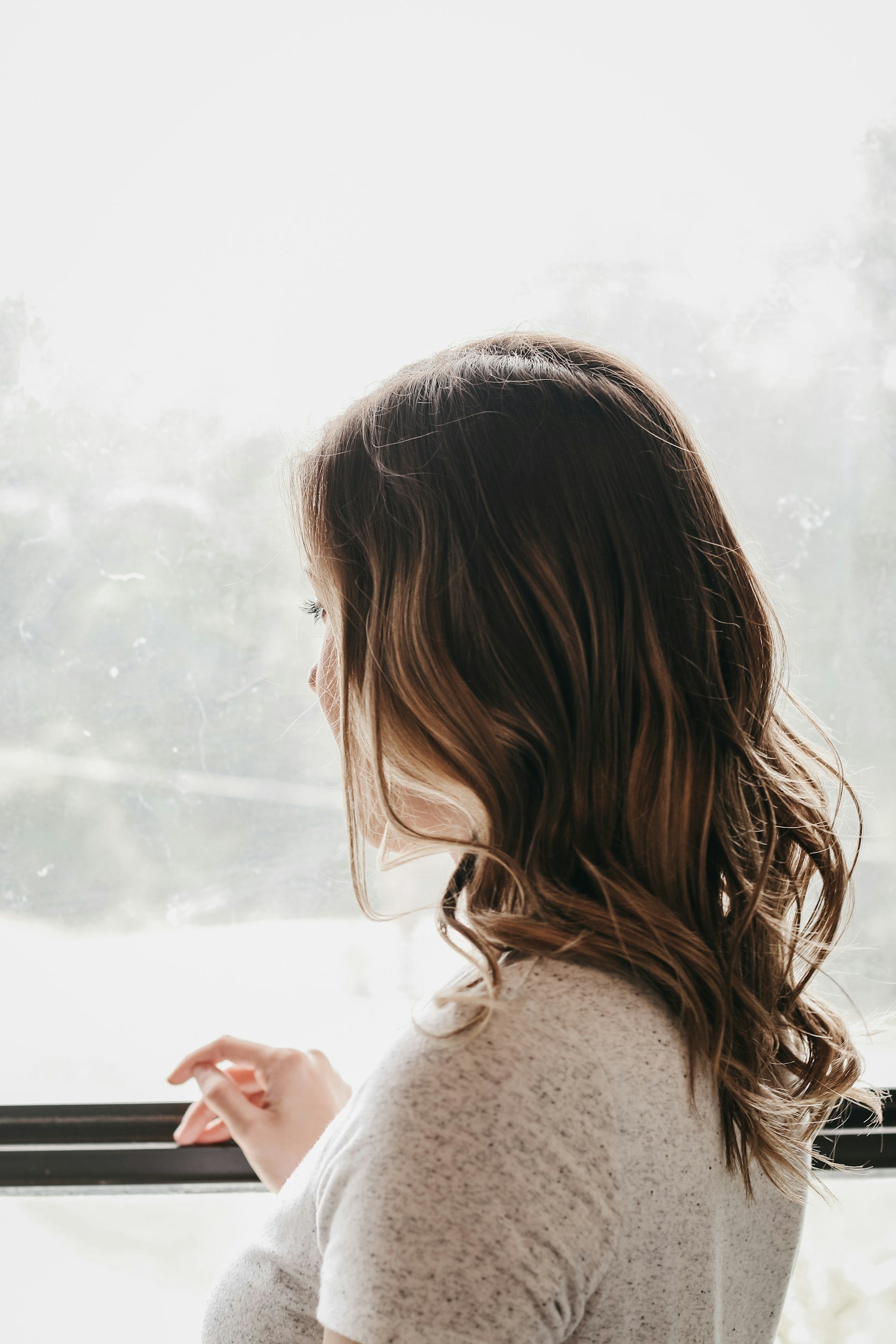 A woman with wavy brown hair looking out a window on a cloudy day, wearing a light-colored sweater and holding a pink cloth in her hand.