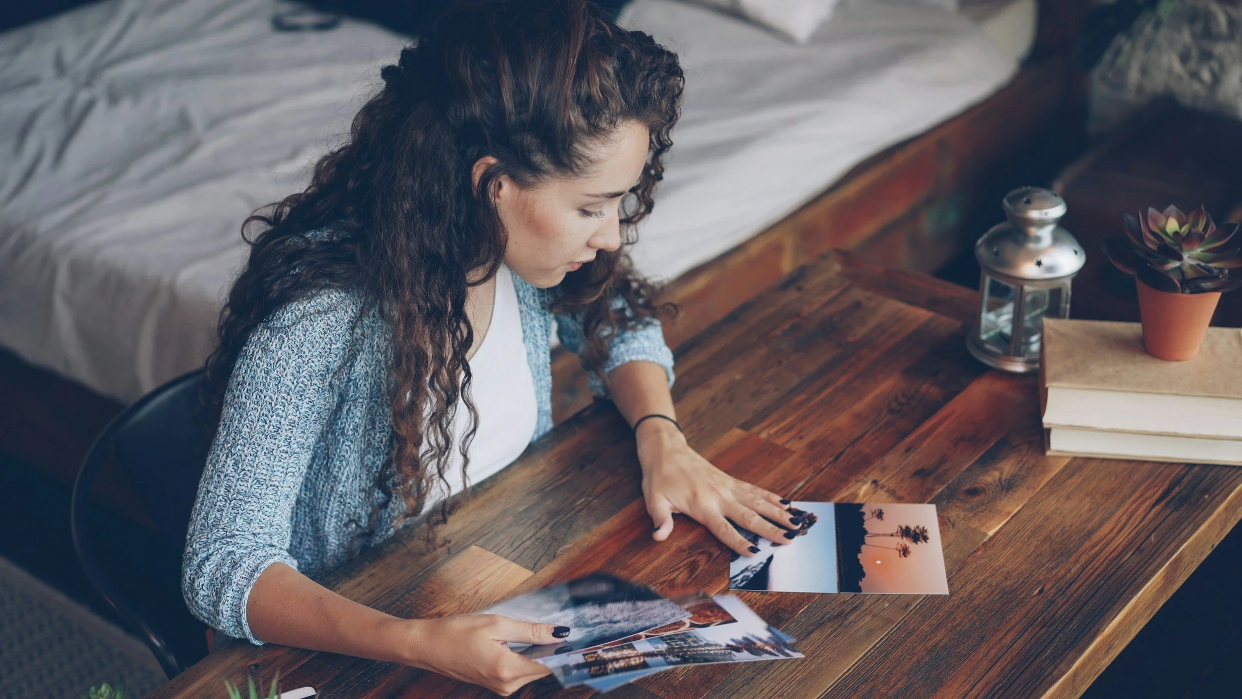 A woman with curly hair sitting at a wooden table, looking at a printed photograph. The table has a small potted succulent, a lantern, and some books.