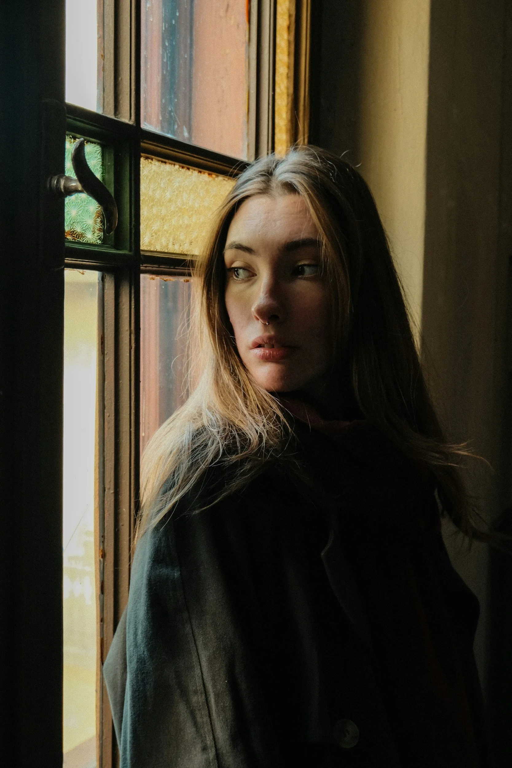A young woman with long dark hair, light-colored eyes, and a septum piercing standing by a window with stained glass. She is wearing a dark hoodie and looking to her right.