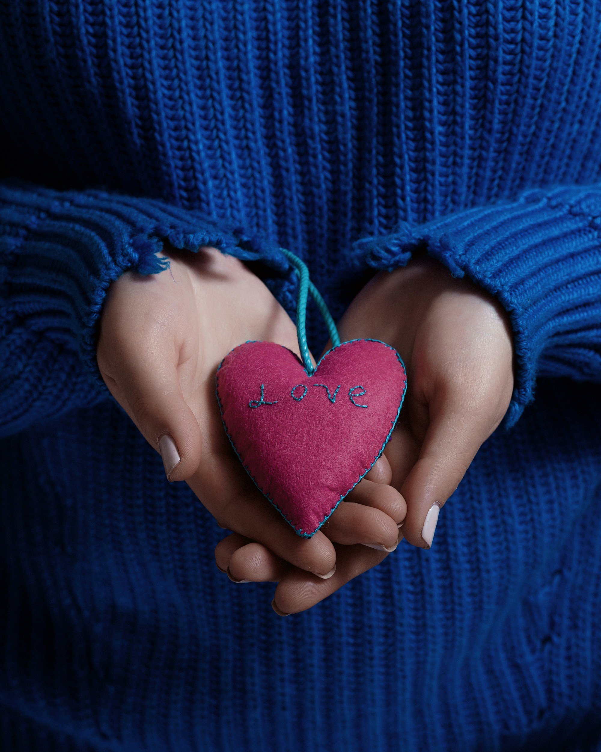 Hands holding a small pink heart with the word "Love" embroidered on it, wearing a blue knitted sweater.