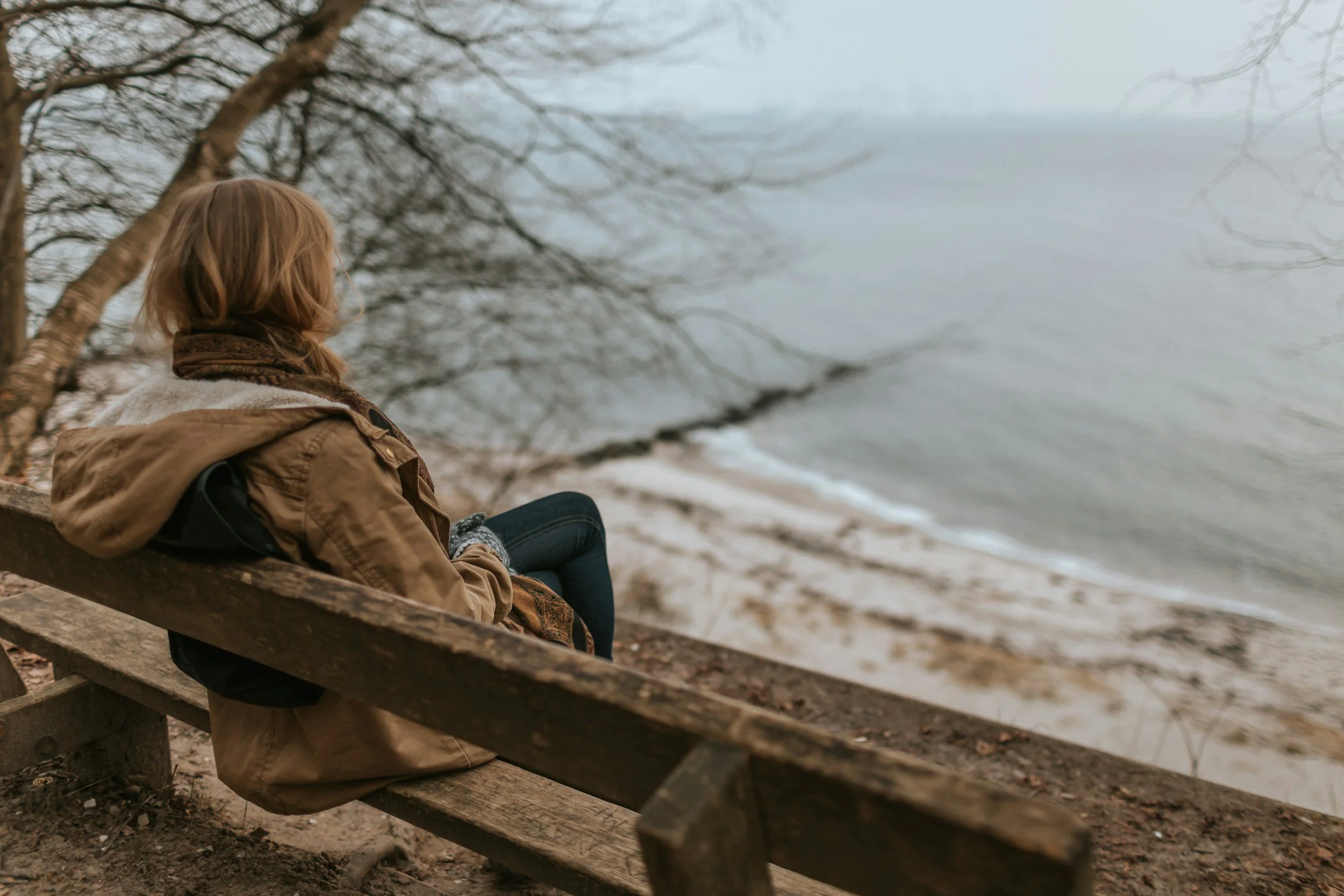 A person with blonde hair, wearing a brown jacket and a scarf, sitting on a wooden bench near a body of water with bare tree branches overhead.