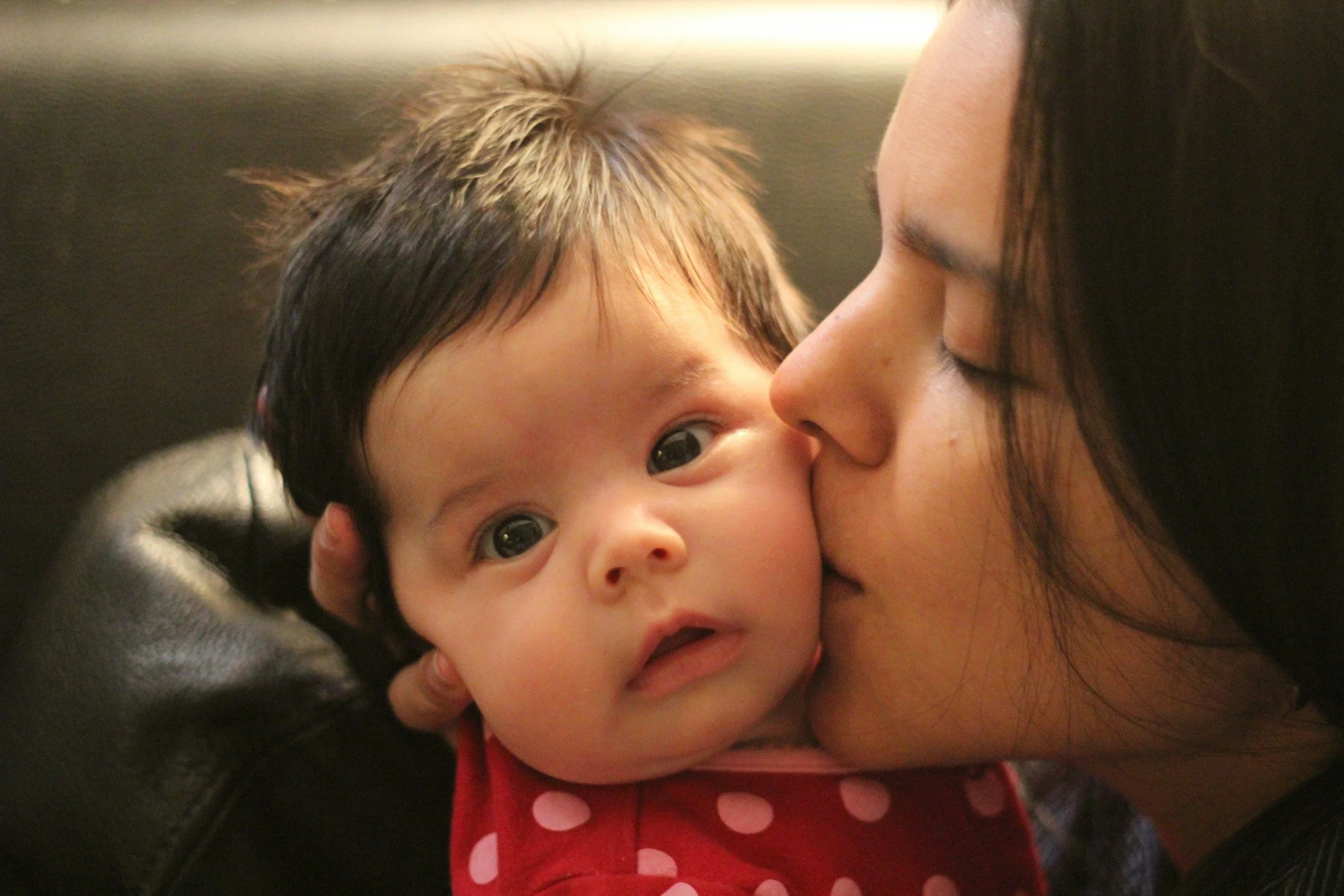 A woman is kissing a young girl on the cheek. The girl has brown hair, big brown eyes, and is wearing a red polka dot dress. The woman has long dark hair and a light brown skin tone.