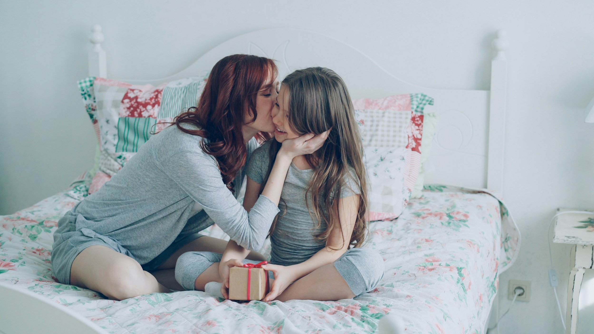 A woman giving a kiss on the lips to a young girl sitting on a bed with floral bedding, holding a small gift box with a red ribbon.