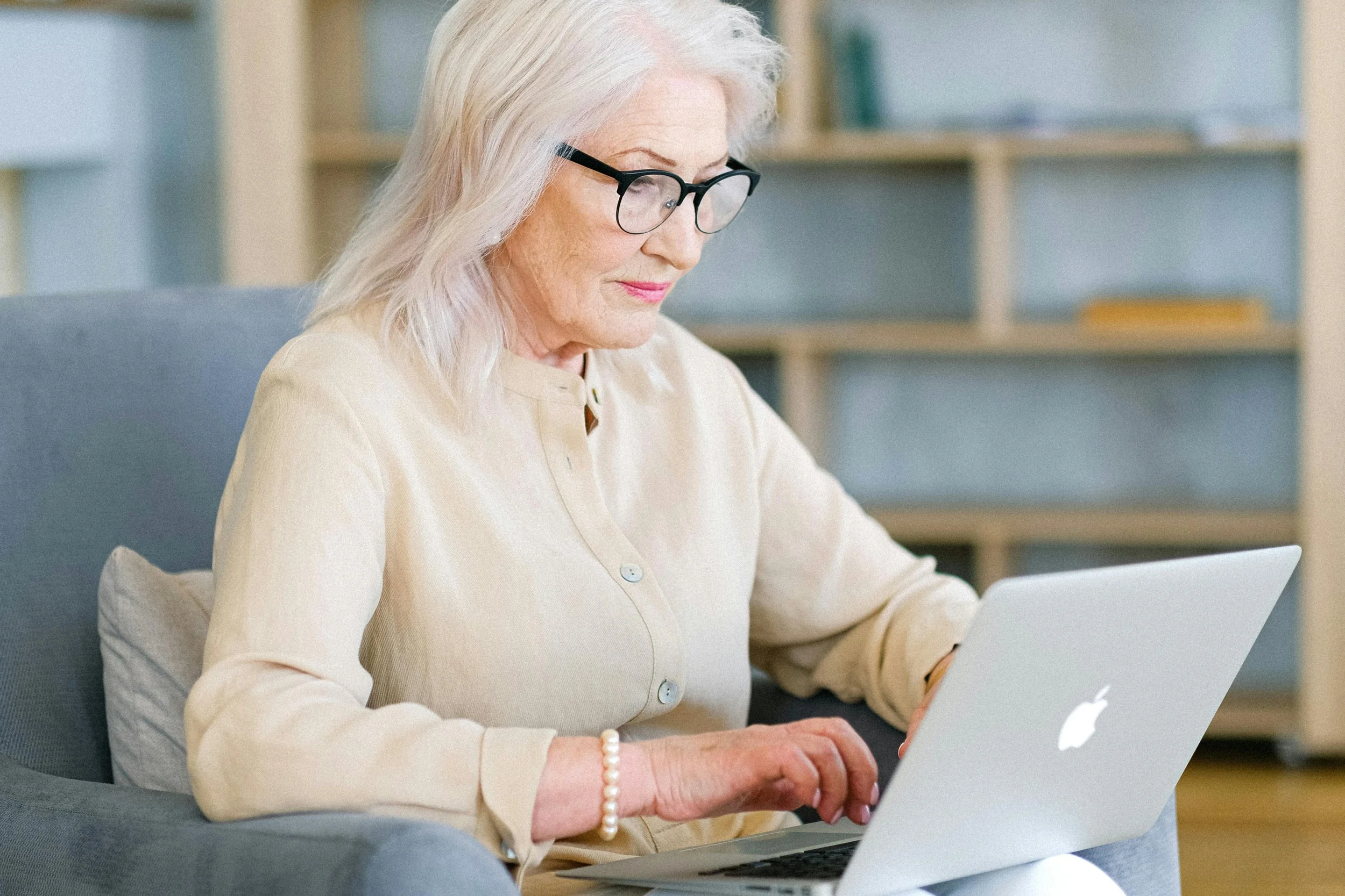 An elderly woman with glasses working on a silver Apple MacBook laptop while sitting on a grey armchair in a room with bookshelves.