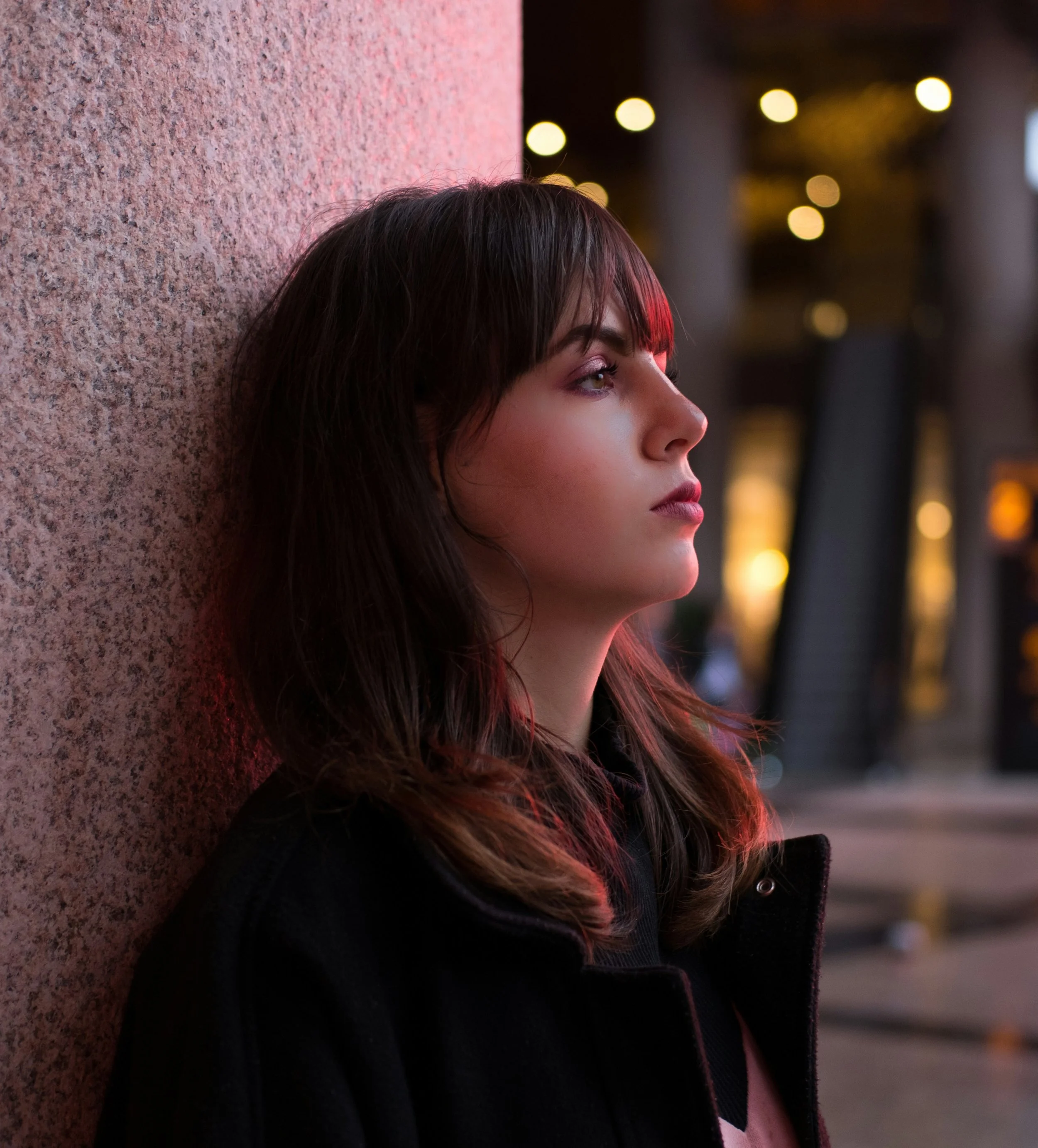 A young woman with dark brown hair and bangs, wearing a black jacket, is sitting against a textured pink wall at night, looking to her right with a calm expression. Blurred city lights are visible in the background.