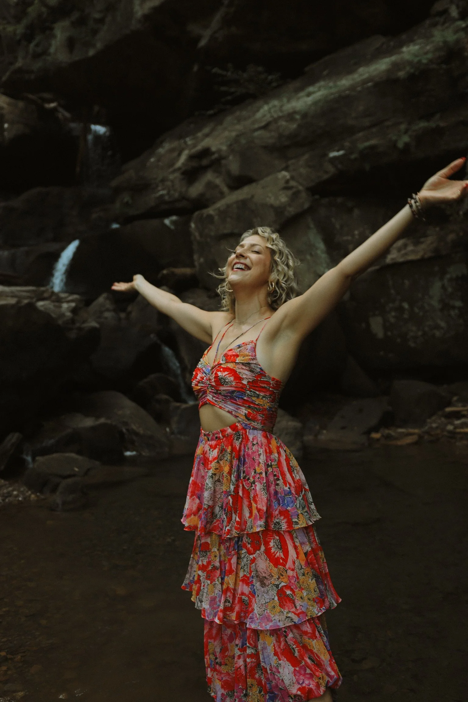 A woman in a colorful, patterned, sleeveless dress with arms outstretched, smiling with her eyes closed, standing in front of a rocky background with small waterfalls.
