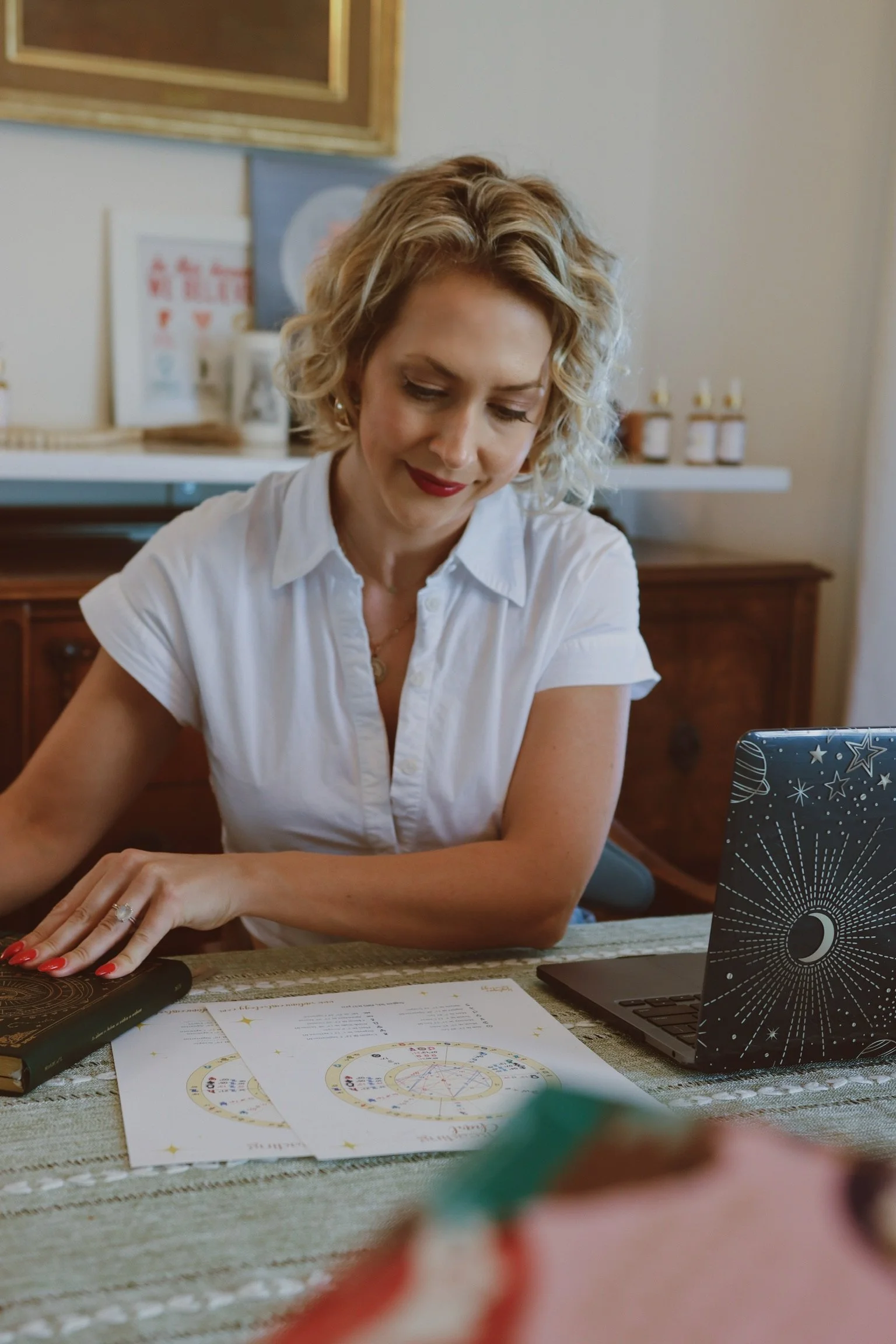 A woman with blonde curly hair, wearing a white shirt, sitting at a table with astrology charts, a black decorated notebook, and a laptop. She is in a room with framed art and shelves with small bottles in the background.