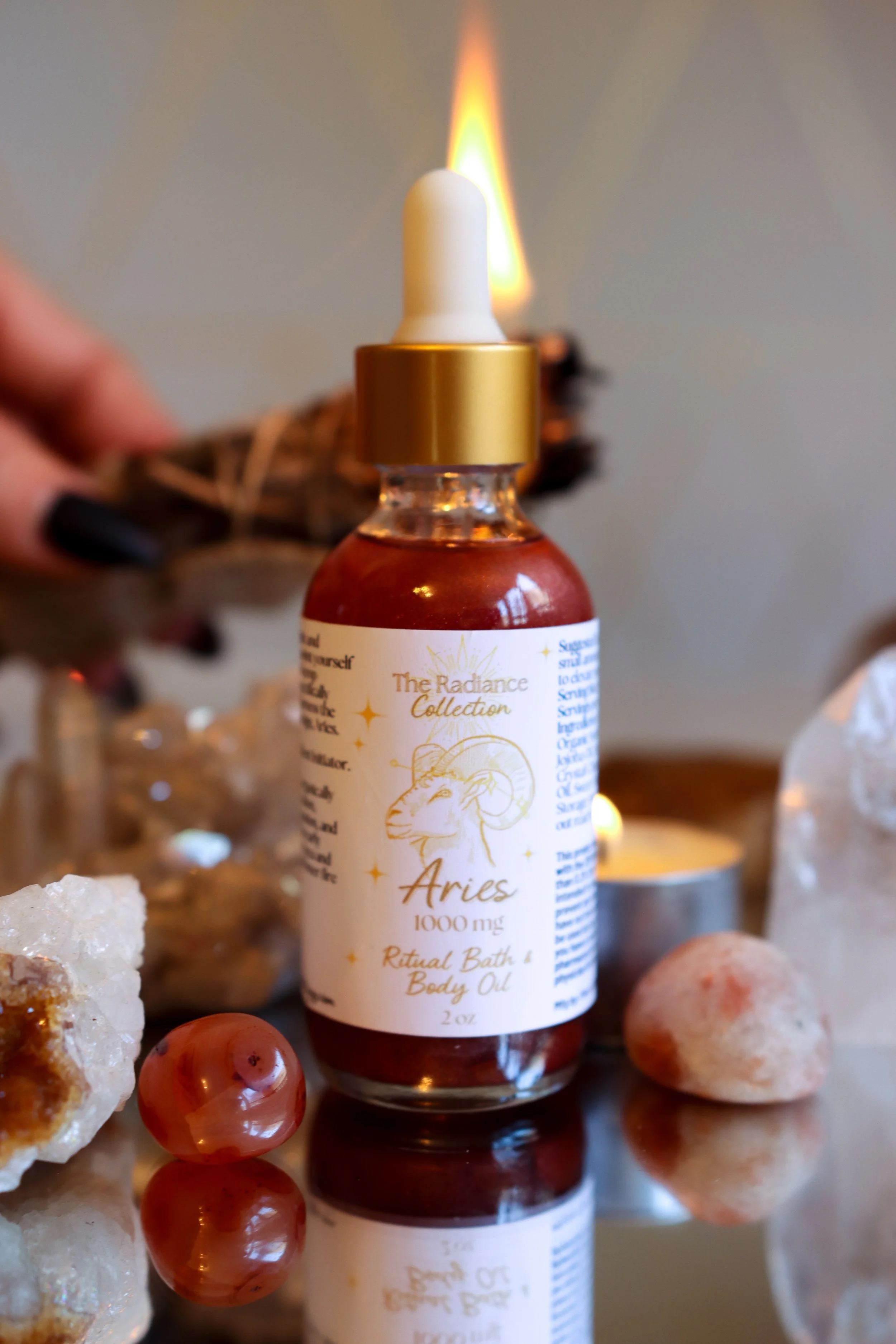 A small bottle labeled 'Aries 1000 mg Ritual Bath & Body Oil' from The Radiance Collection, with a lit candle, colored stones, and crystals around it on a reflective surface.