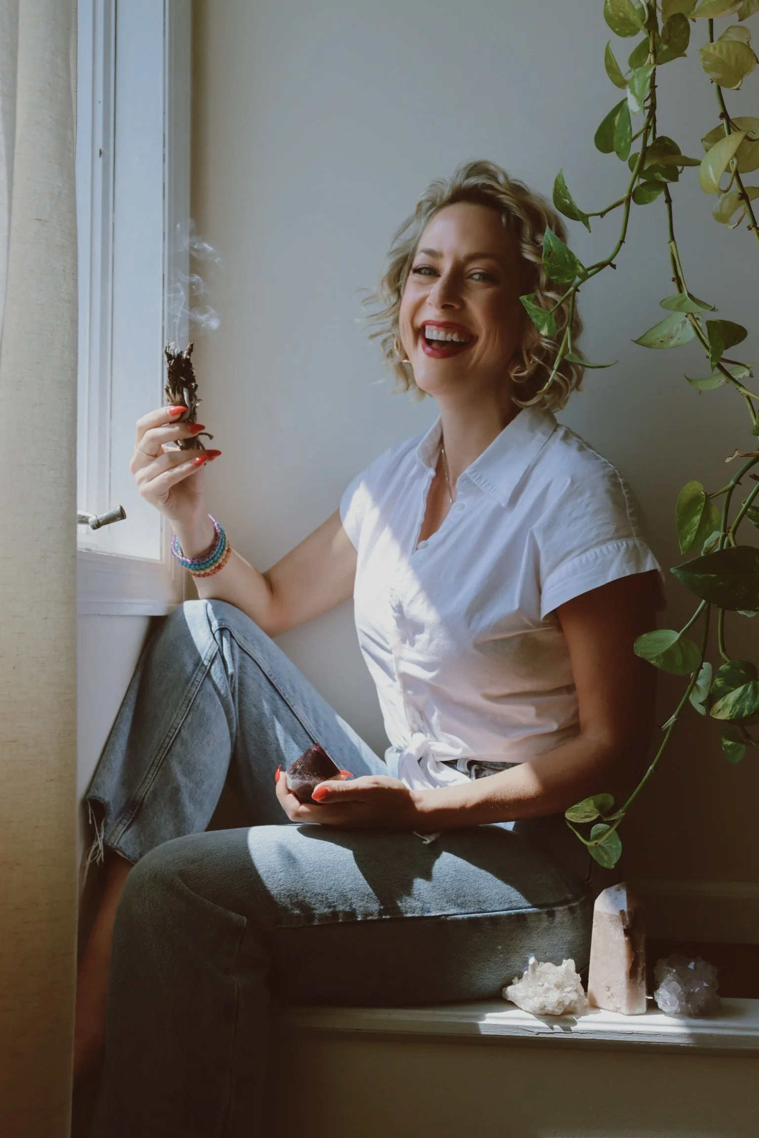 A woman with curly blond hair, wearing a white shirt and blue jeans, sitting by a window with sunlight. She is smiling and holding a lit cigarette or joint in one hand and a small object in the other. There is a leafy plant in the background and crystals on the window sill.