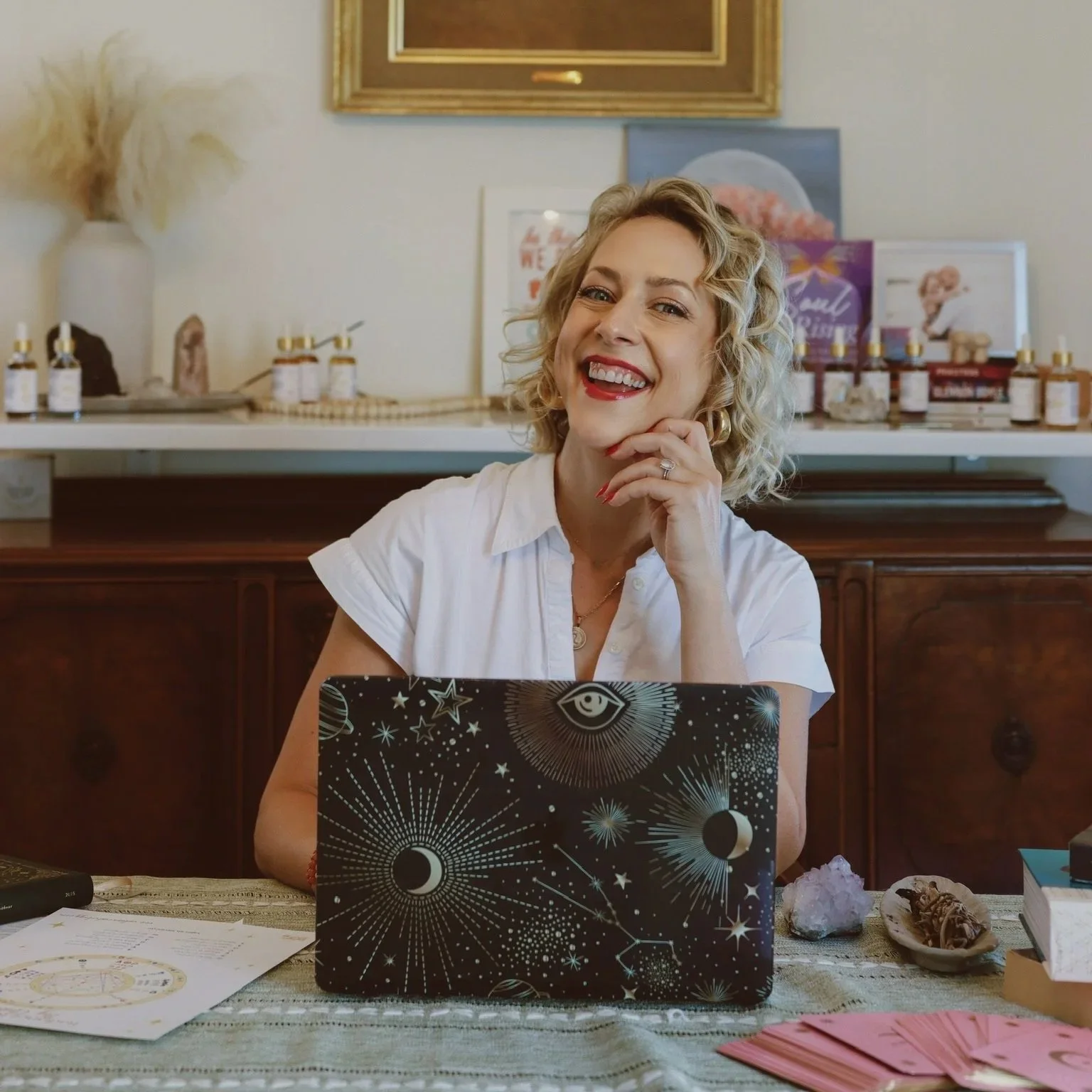A smiling woman with curly blonde hair and red lipstick sitting at a table with a laptop in front of her, surrounded by various objects including crystals, papers, and decorative items in a cozy indoor space.