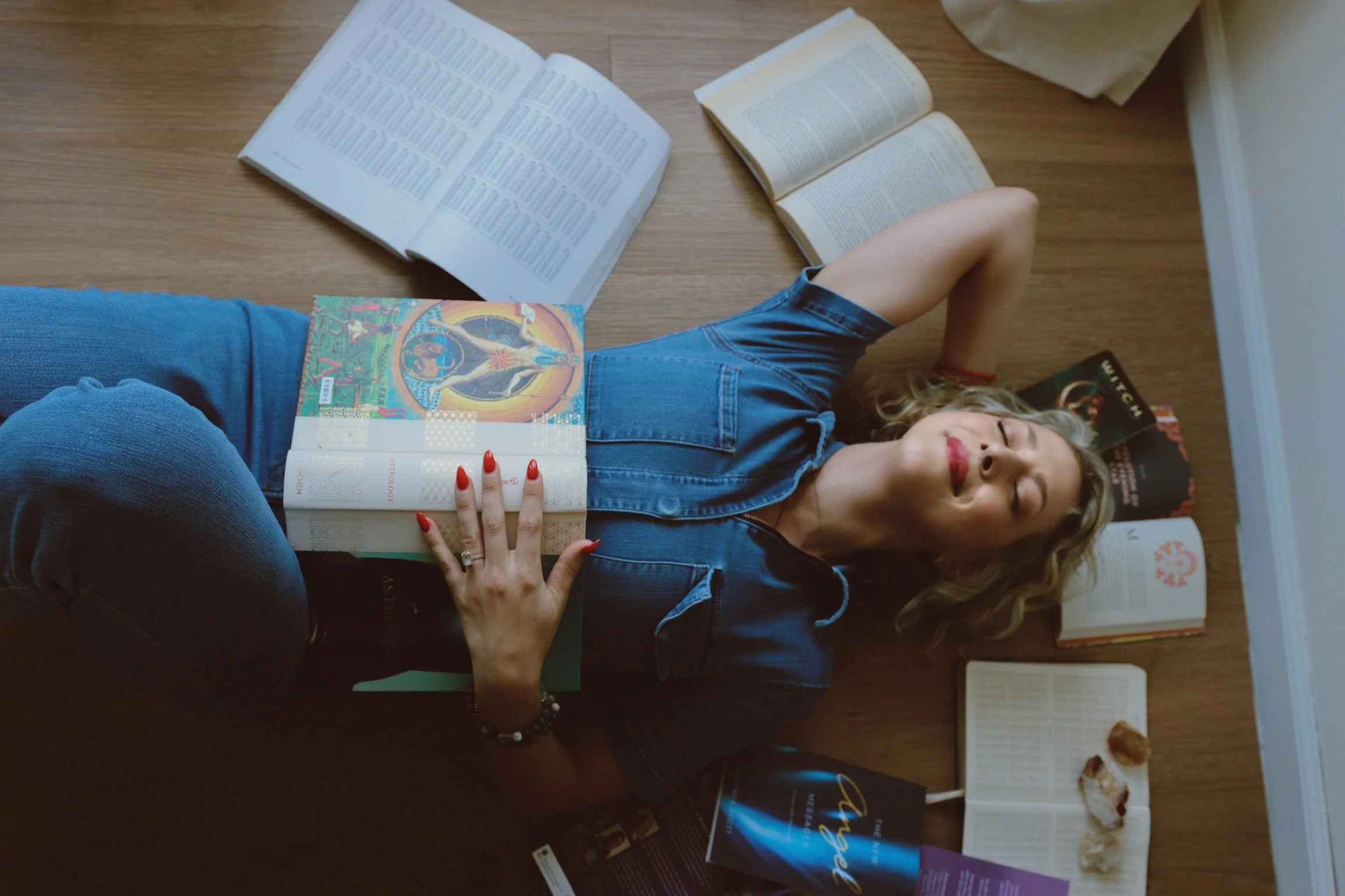 A woman lying on her back on a wooden floor, surrounded by open books and reading materials, with her eyes closed and blonde curly hair spread out around her.