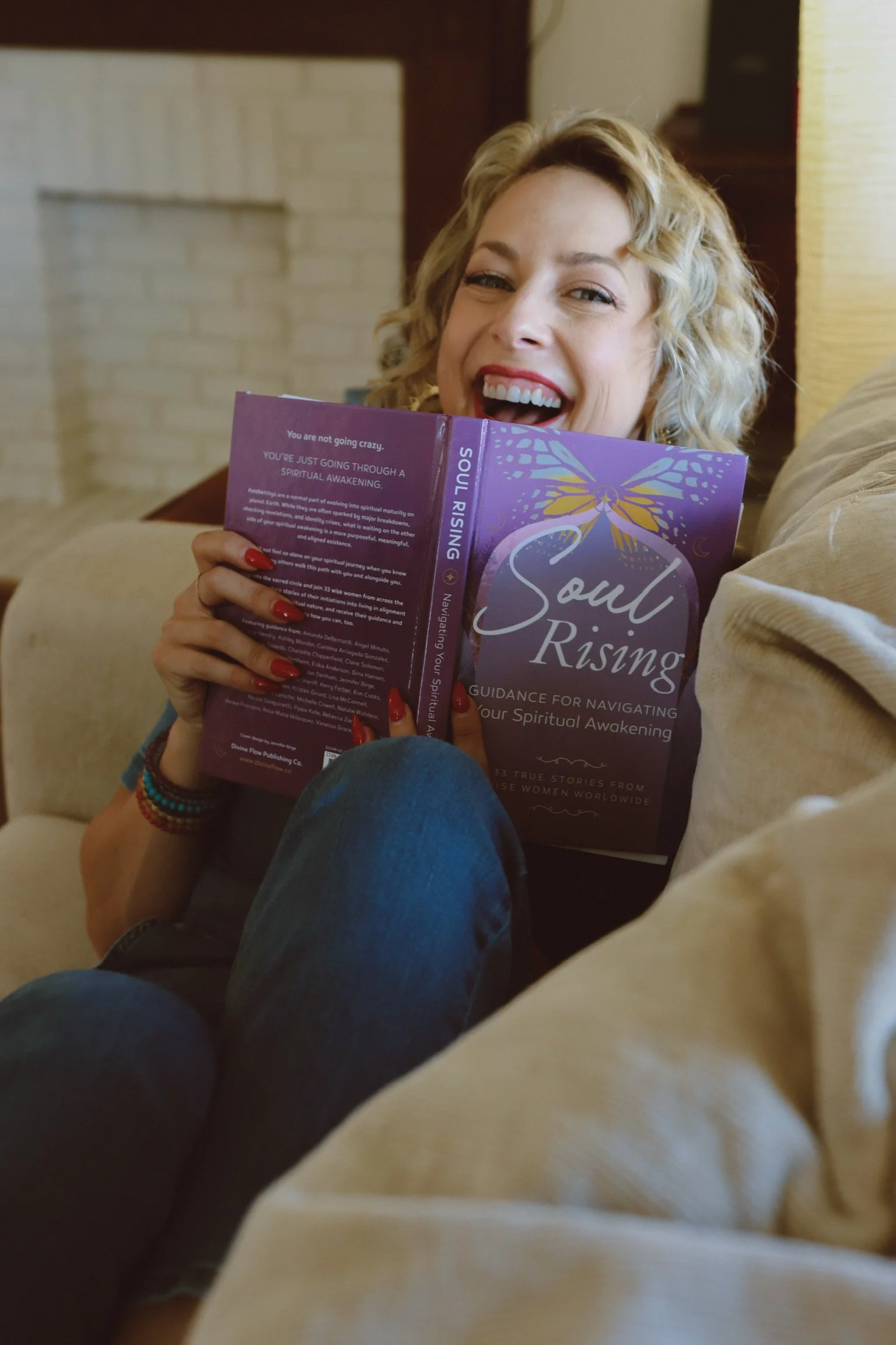 A woman with curly blonde hair sitting on a beige couch, smiling and holding a purple book titled "Soul Rising." She is wearing a black shirt and red nail polish.
