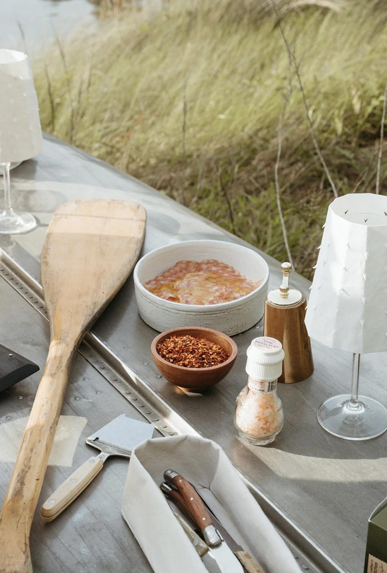 Outdoor table set with a pizza, spices, salt, pepper grinder, wine glasses, and utensils by a grassy field.
