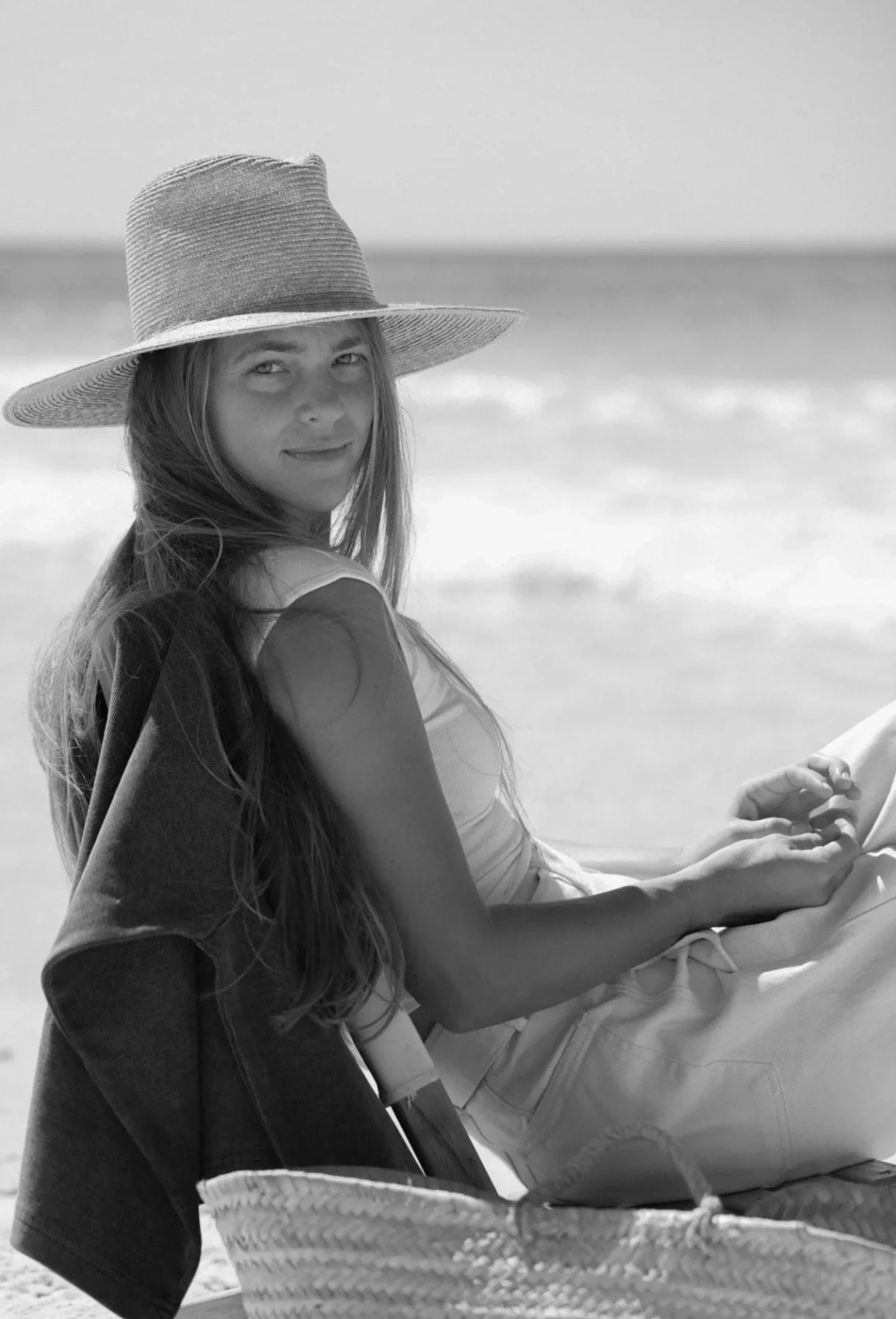 A woman in a large sunhat and sleeveless top sitting on a beach with waves in the background.