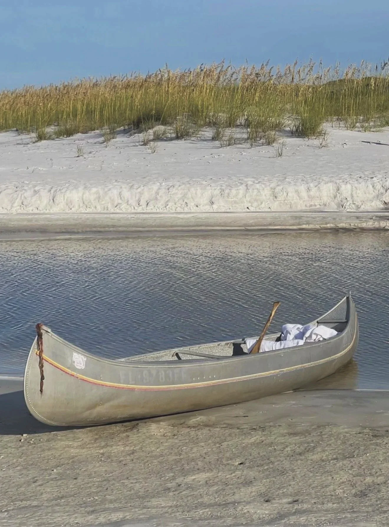 A small canoe resting on the sandy shore of a calm body of water with white sand dunes and tall grass in the background under a clear sky.
