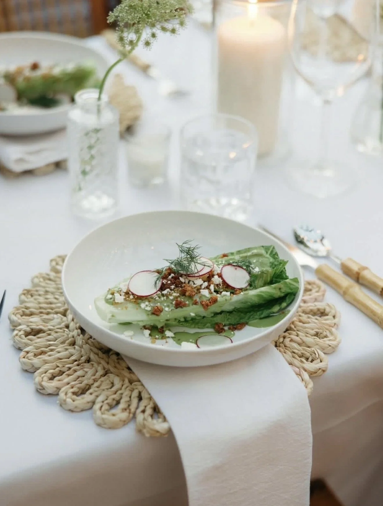 A white plate with salad garnished with radish slices, herbs, and crumbled cheese on a table with a woven placemat, surrounded by glasses, candles, and a vase with flowers.