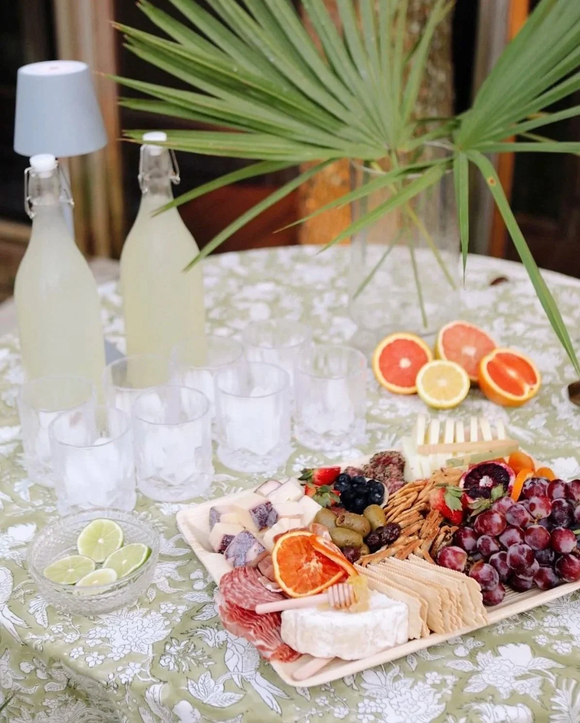 A table set with a platter of assorted cheeses, meats, grapes, crackers, and fruit slices, with glasses, lemonade bottles, and lemon and grapefruit halves, decorated with large green palm leaves on a floral tablecloth.