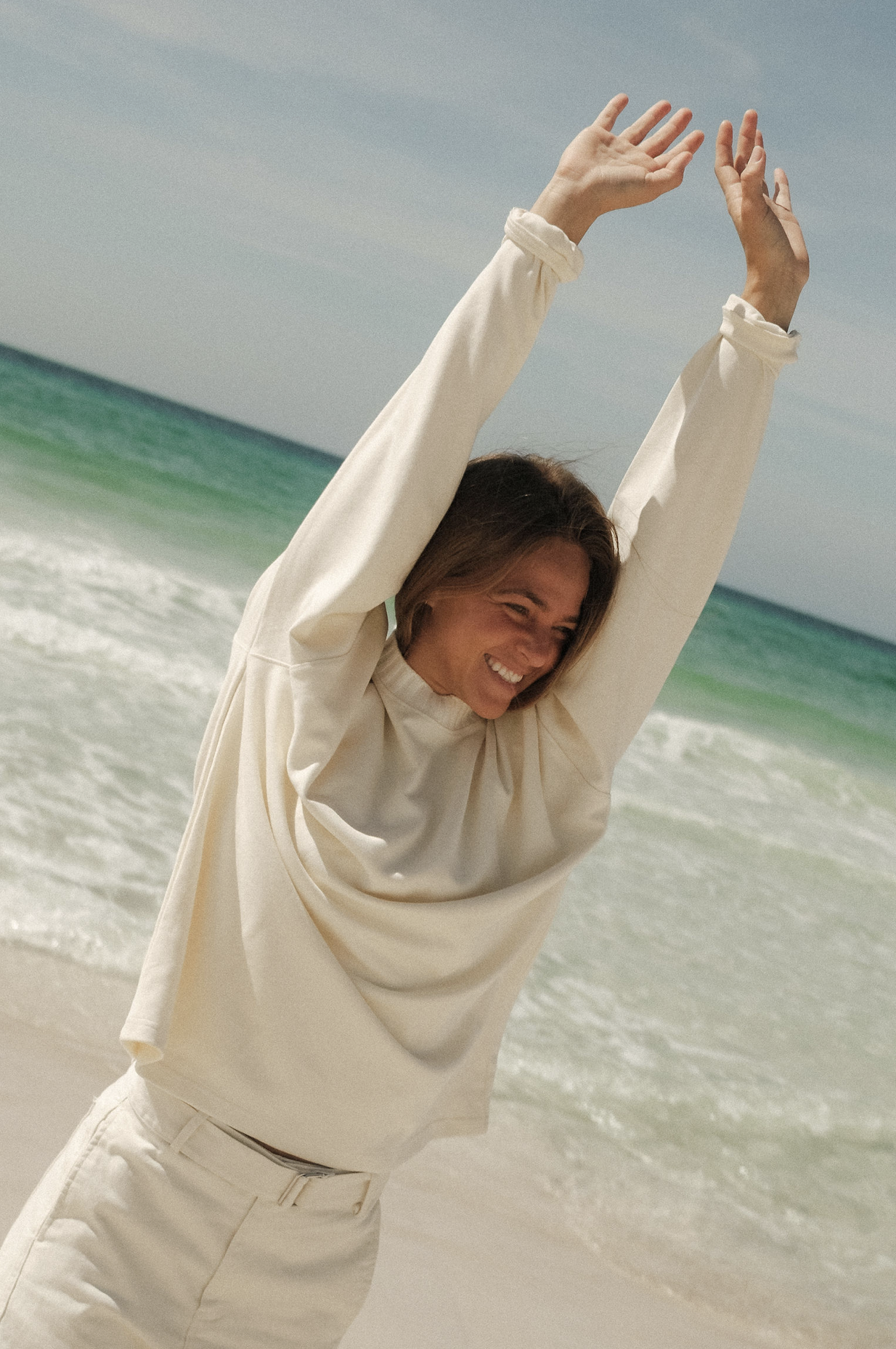 A woman in a cream-colored outfit joyfully stretching on a beach with the ocean in the background.