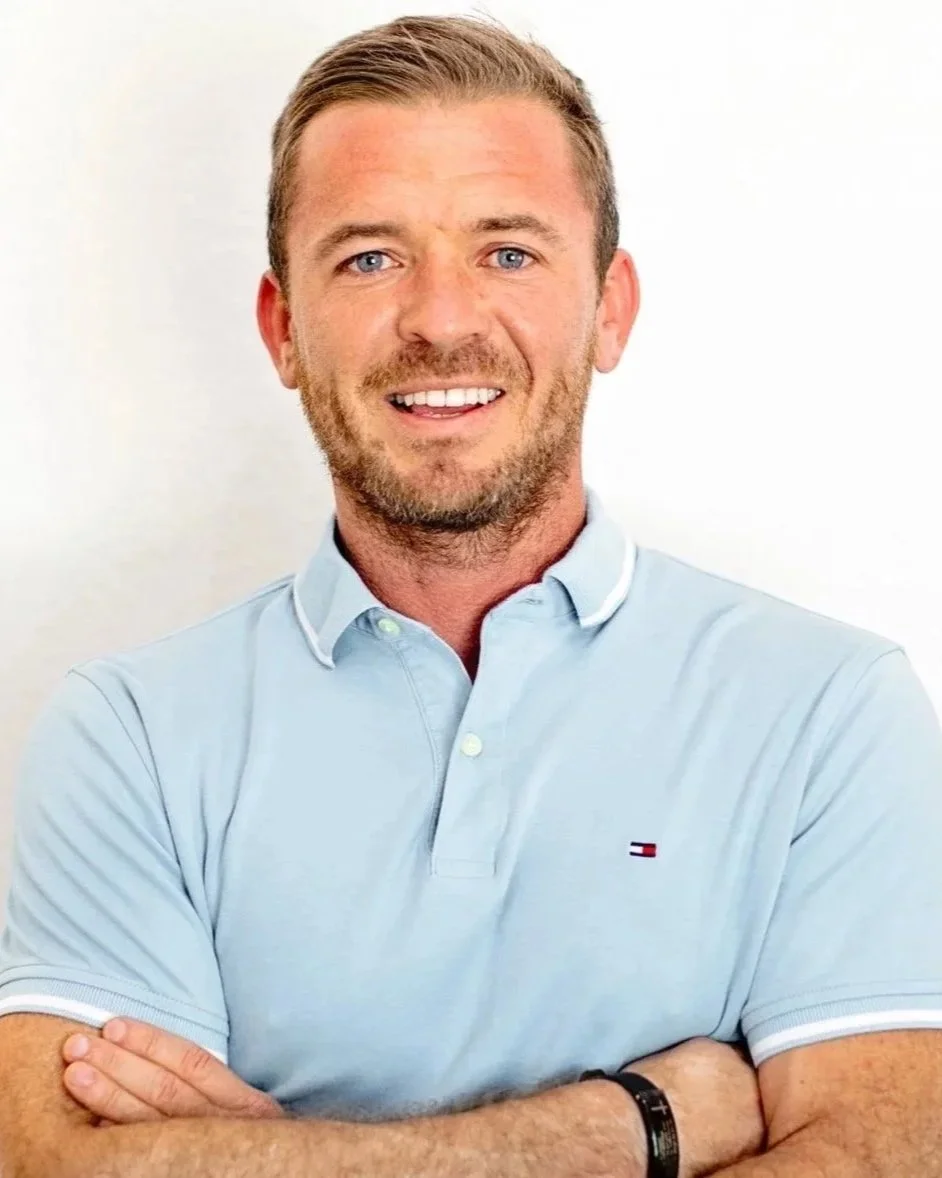 A smiling man with blue eyes, short light brown hair, and a beard, wearing a light blue polo shirt with white trim, standing with arms crossed against a plain white background.