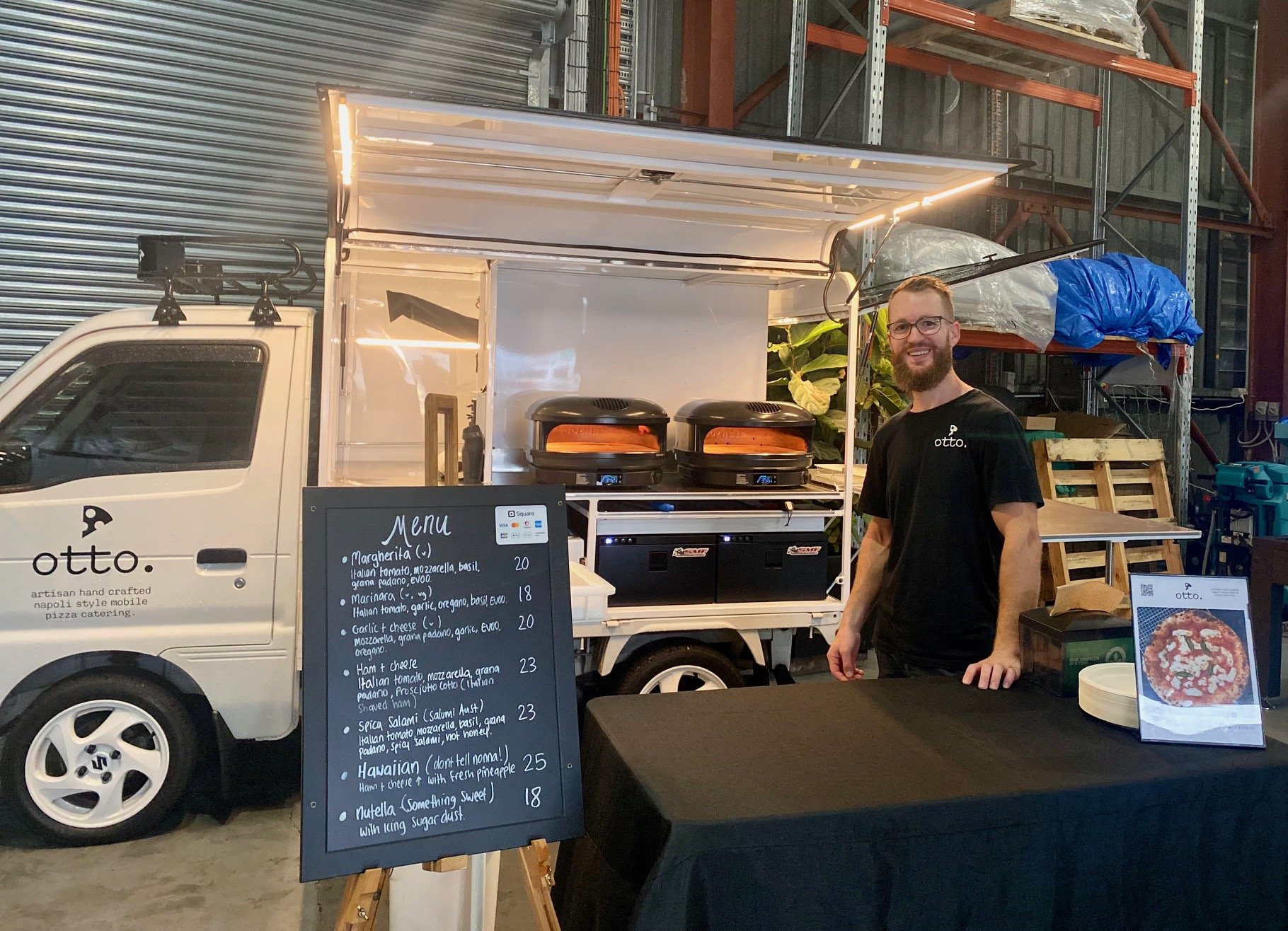 Mobile pizza catering setup with a small white truck featuring an open kitchen area, displaying a menu board and cooking equipment. A man is standing beside the setup smiling, wearing a black t-shirt with 'otto.' written on it. The menu board lists v