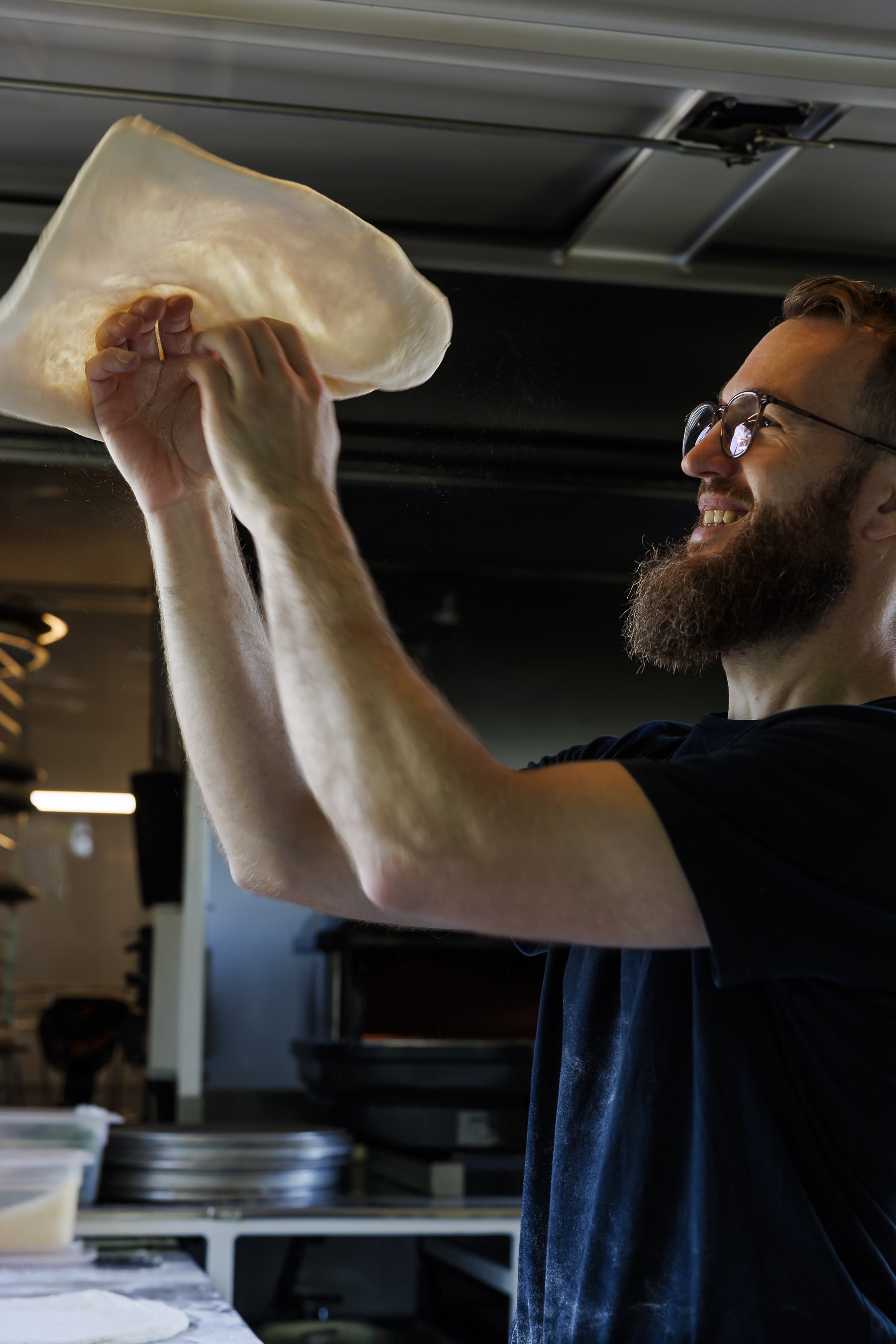 A man with glasses and a beard is in a kitchen, stretching pizza dough with his hands.