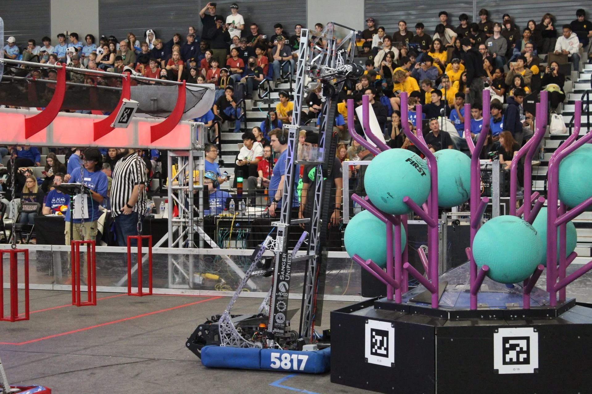 A robotics competition with a robot preparing to shoot balls into a target, a group of spectators in the stands, and a large green and purple ball intake system in the foreground.