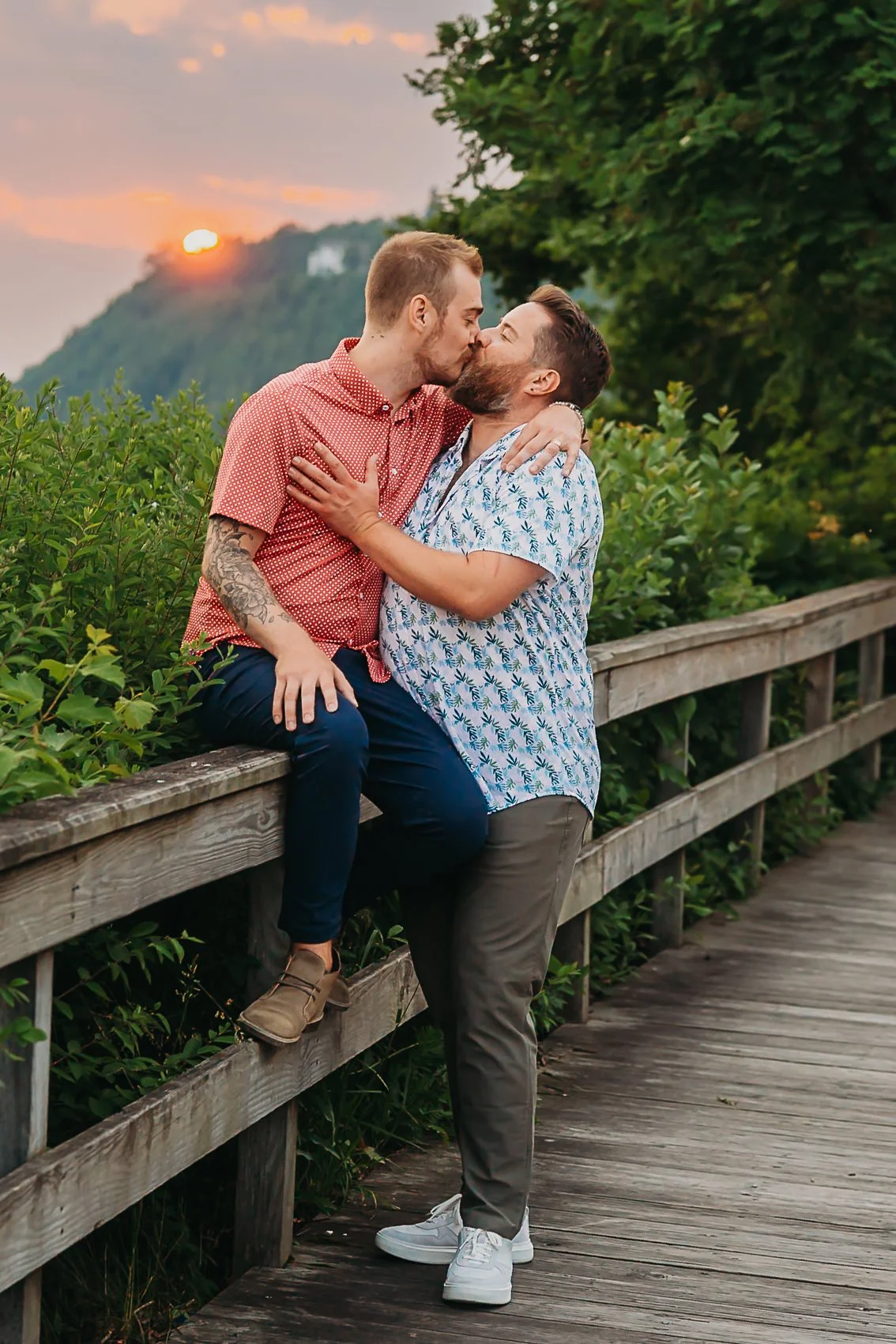 Two men sharing a kiss on a wooden walkway with greenery and a sunset in the background on Mackinac Island