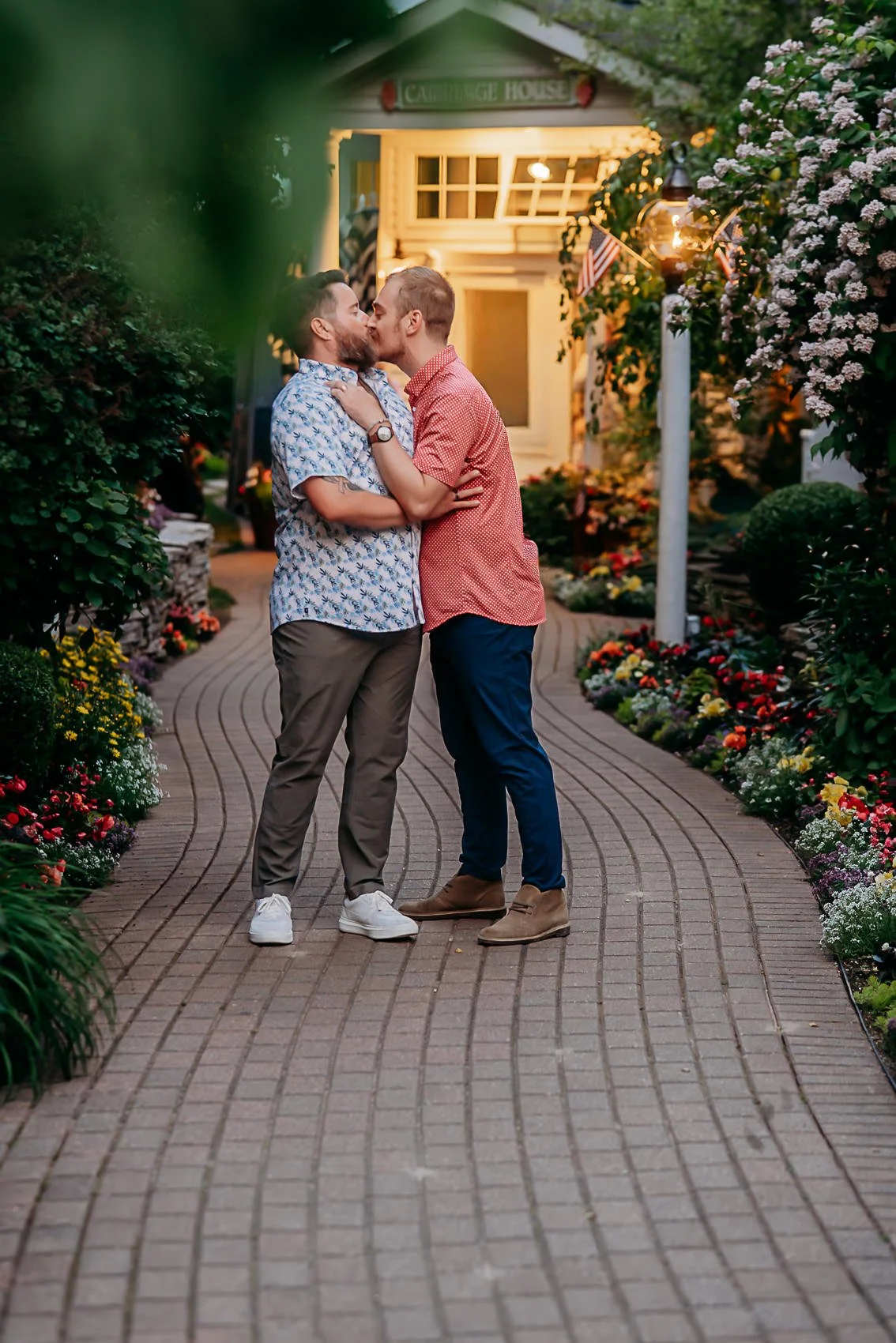 Recently engaged couple sharing a kiss on a flower-lined brick path in front of the Carriage House at dusk.