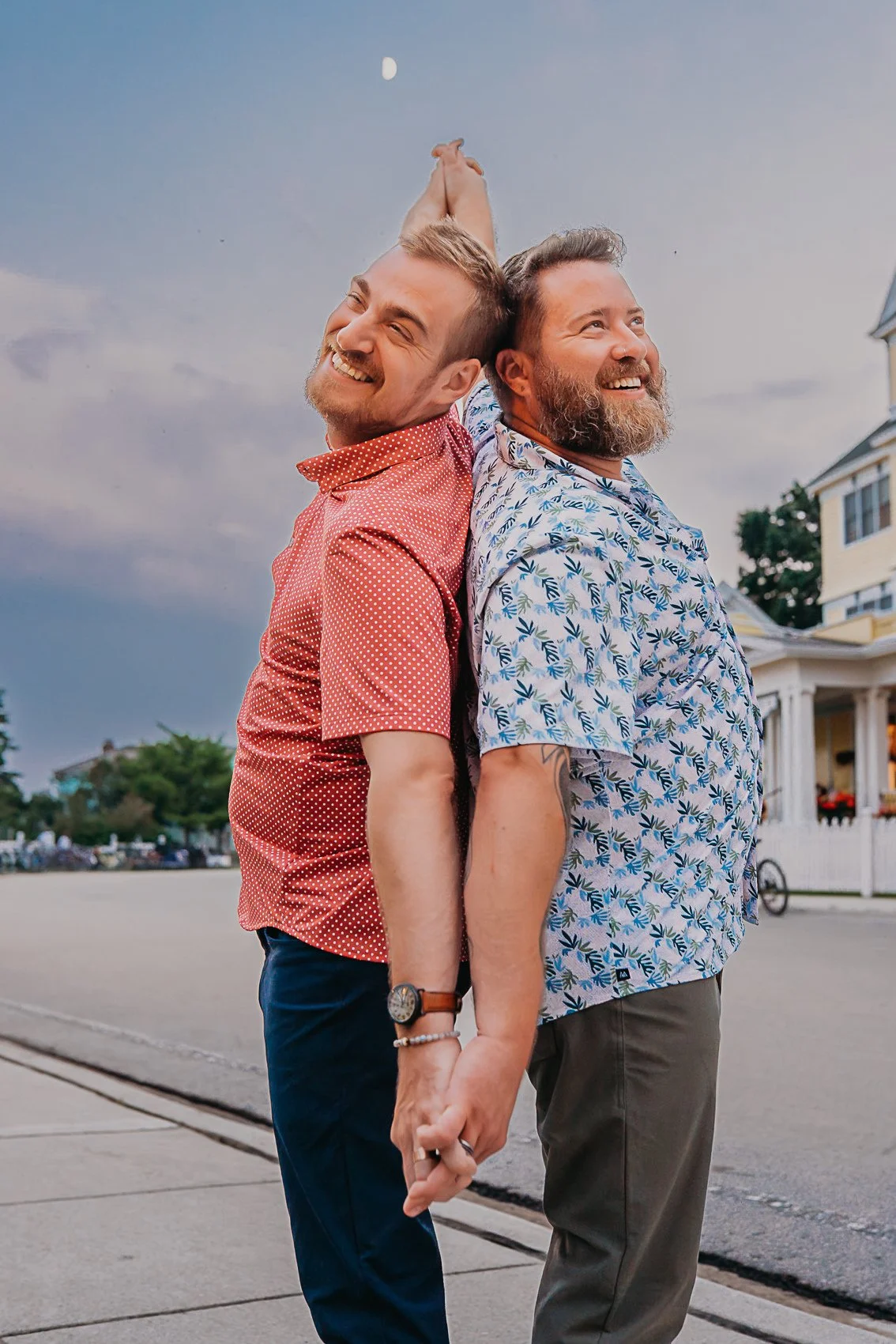 Two smiling men standing back-to-back, holding hands, outdoors in front of a house, with the moon visible in the sky.